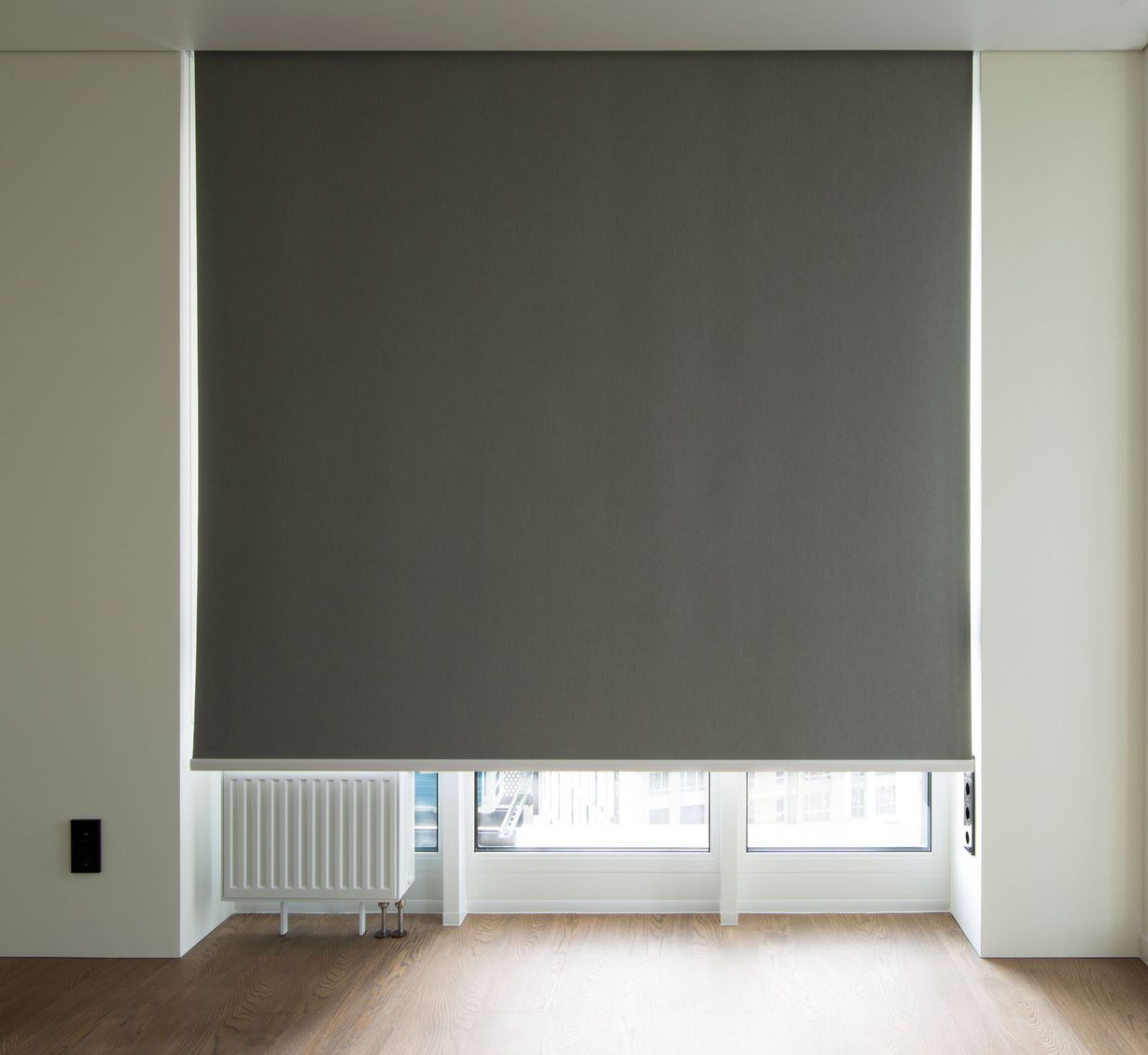 Empty room with a large gray roller blind covering a window, light wood floor, and a radiator below.