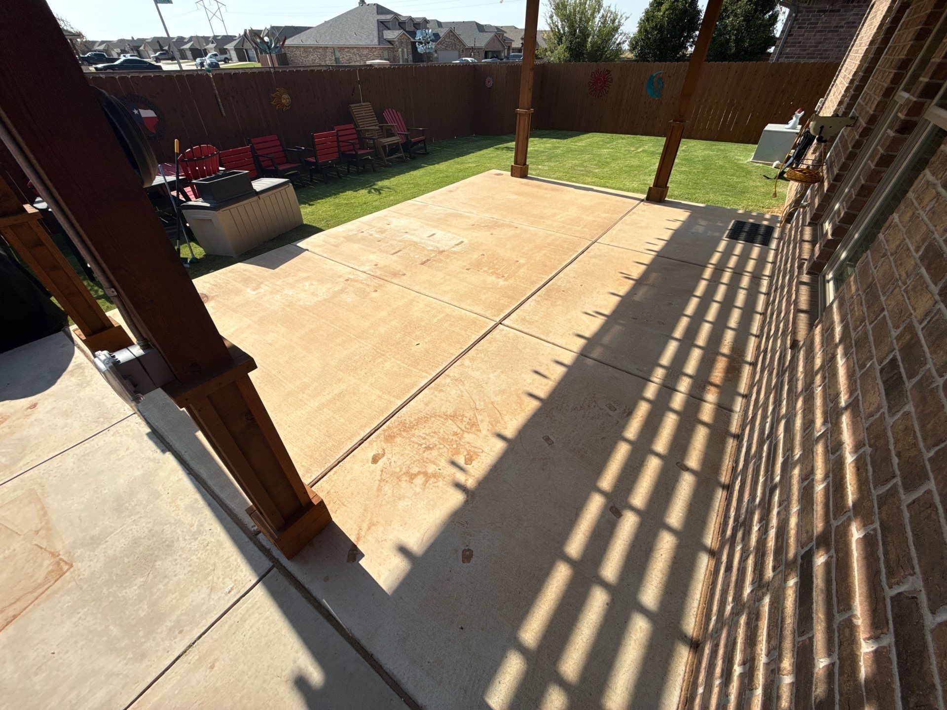 Sunny backyard patio with tan concrete, wooden posts, green lawn, and striped shadows from a railing
