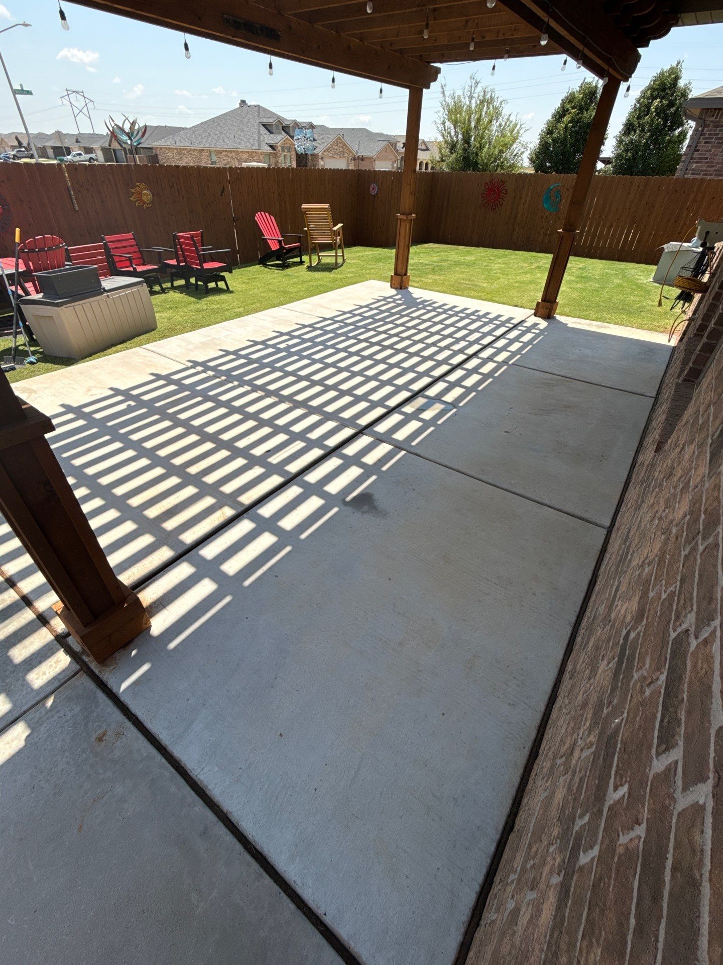 Covered backyard patio with concrete slab, lawn, red chairs, and striped shadows from the pergola.