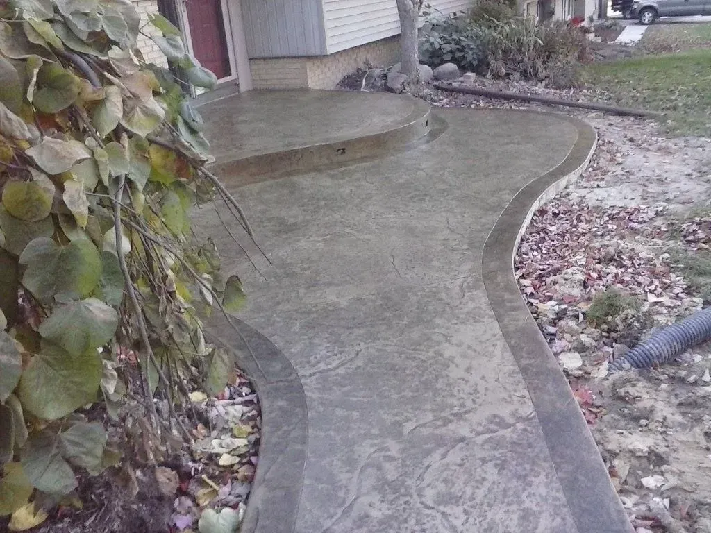 A concrete walkway leading to a house with leaves on the ground