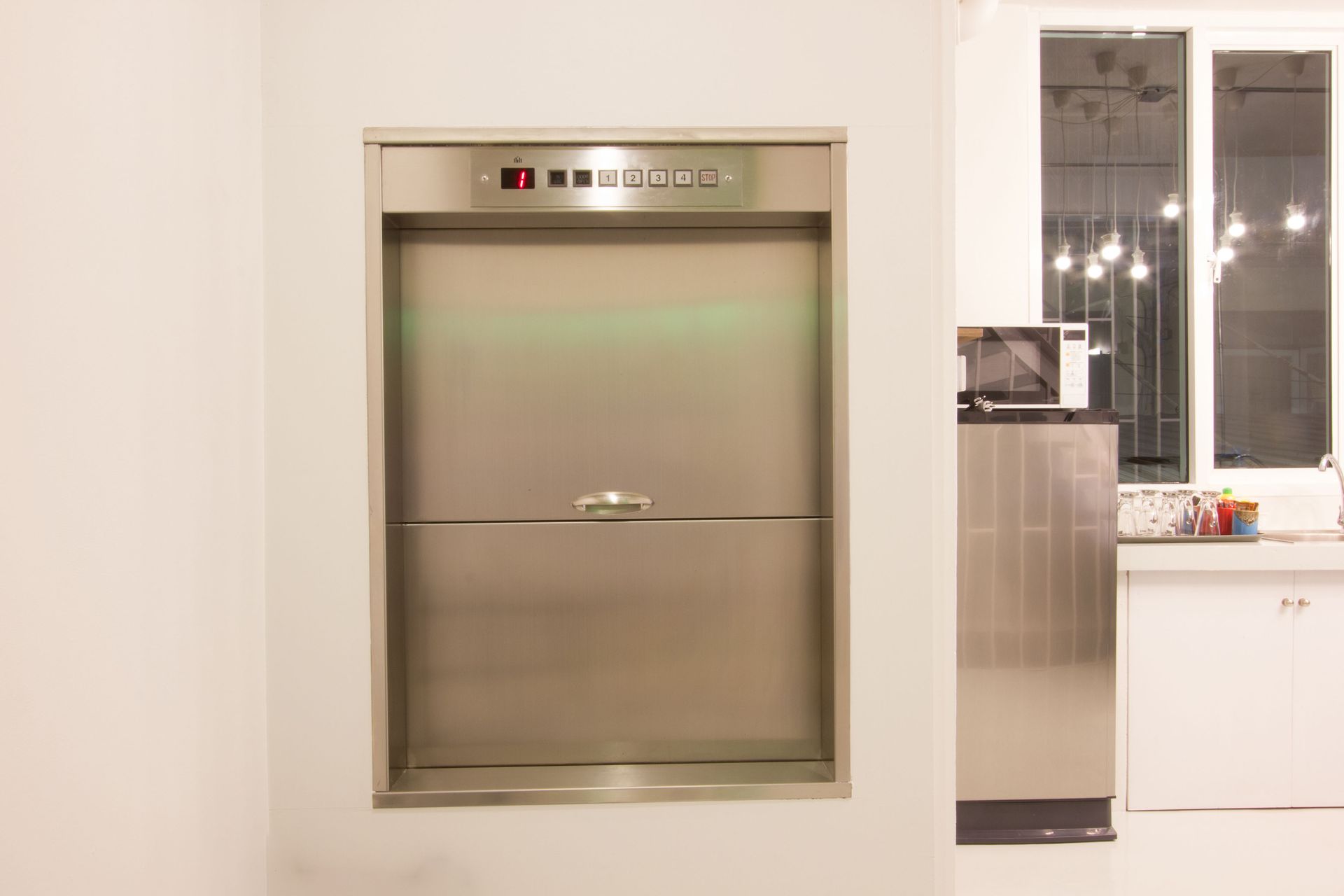 Stainless steel dumbwaiter built into a white wall; kitchen visible in background.