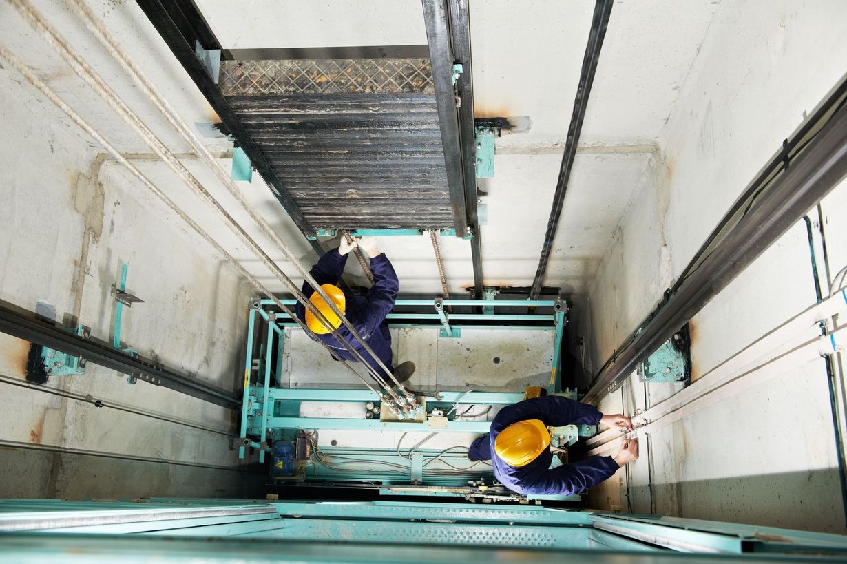 Two elevator technicians in yellow helmets work inside an elevator shaft.