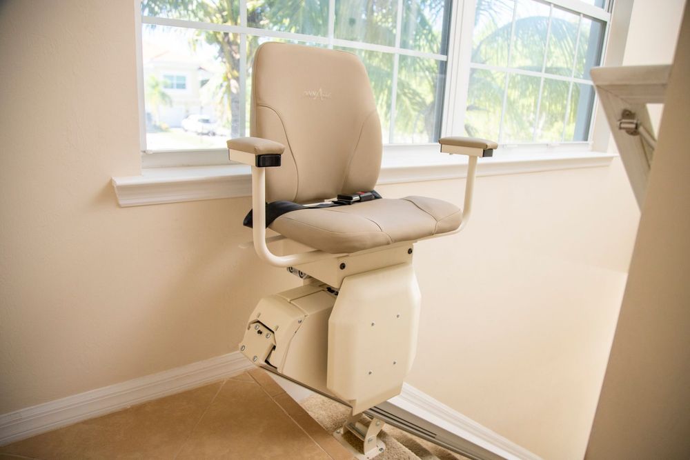 Beige stairlift chair by a window on a light-colored wall.