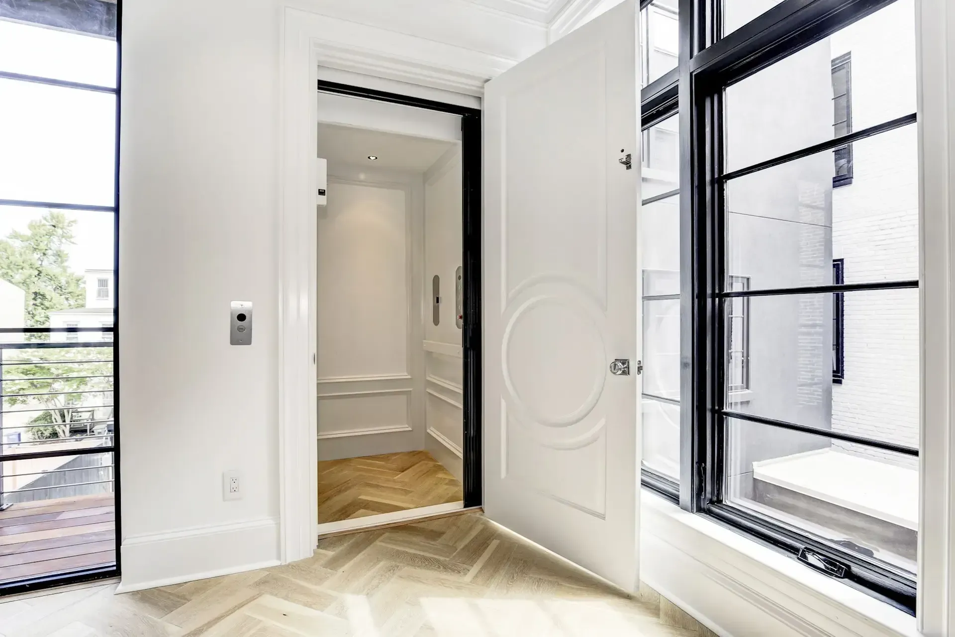 An interior view of a white elevator with an open door, next to windows with black frames.