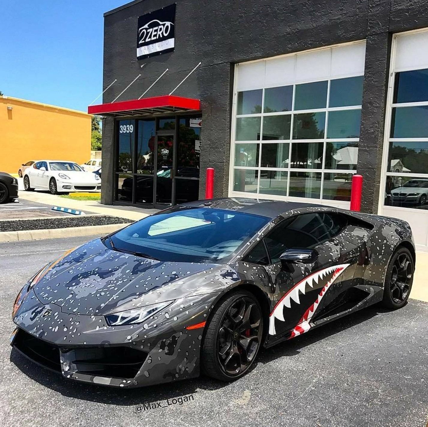 Lamborghini with grey camouflage wrap and shark mouth graphic parked outside a business with a black facade.