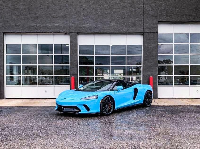 Bright blue McLaren sports car parked in front of a gray building with white garage doors.
