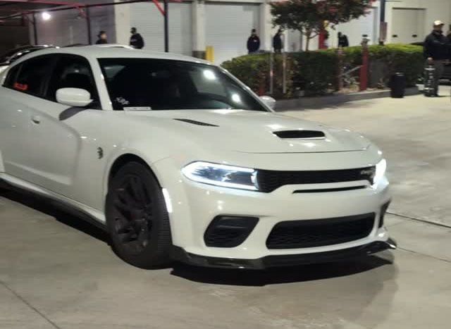 White Dodge Charger with black wheels and accents, front view. Nighttime setting with people in background.