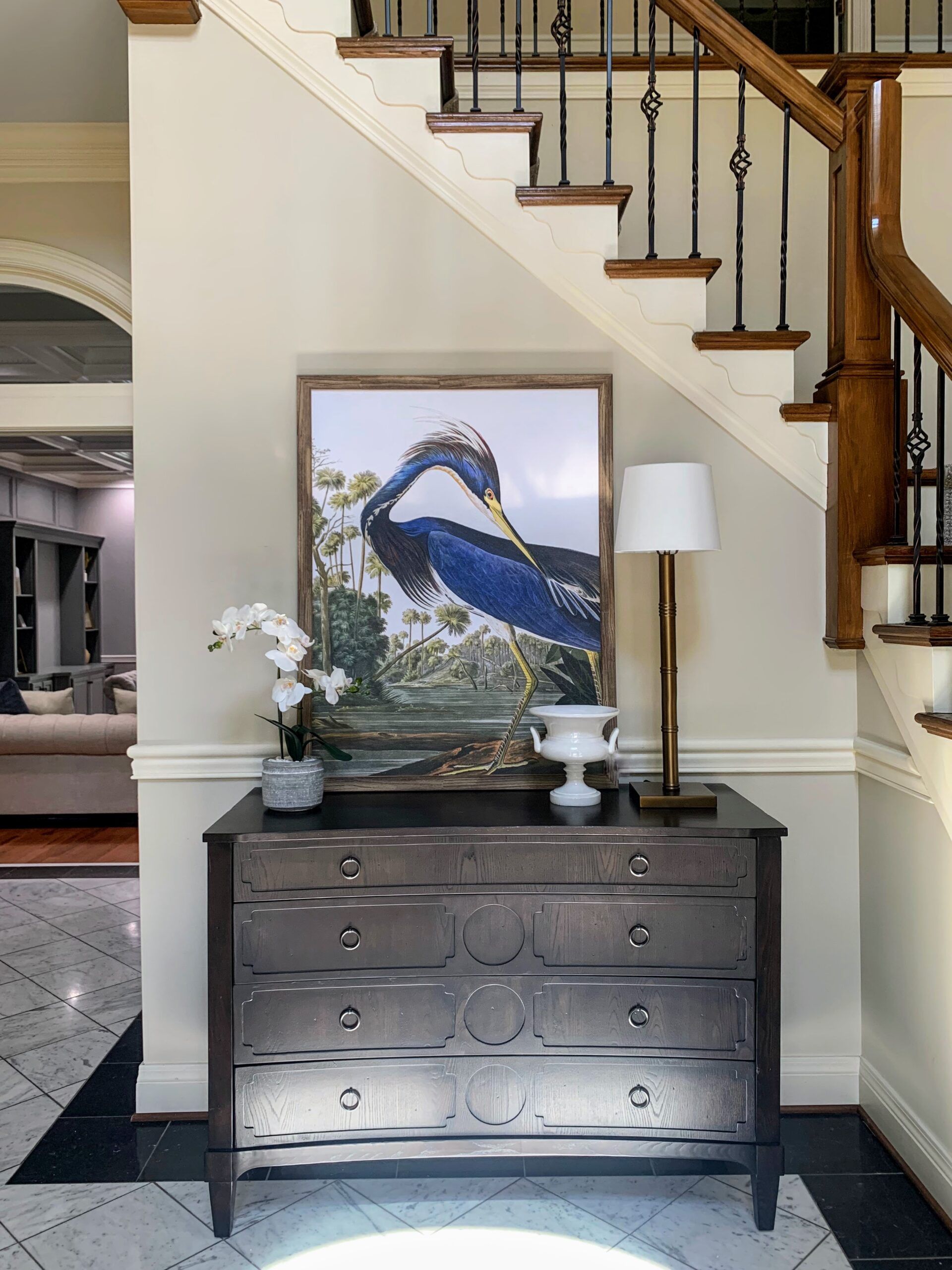 Dark wood dresser with bird painting, lamp, and flowers beneath a staircase in a home interior.
