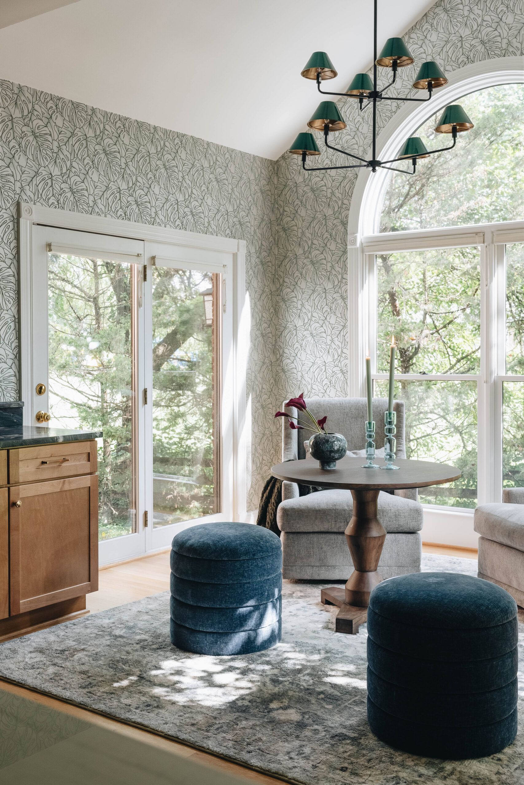 Bright dining nook with patterned wallpaper, round table, blue stools, and large arched windows overlooking greenery.