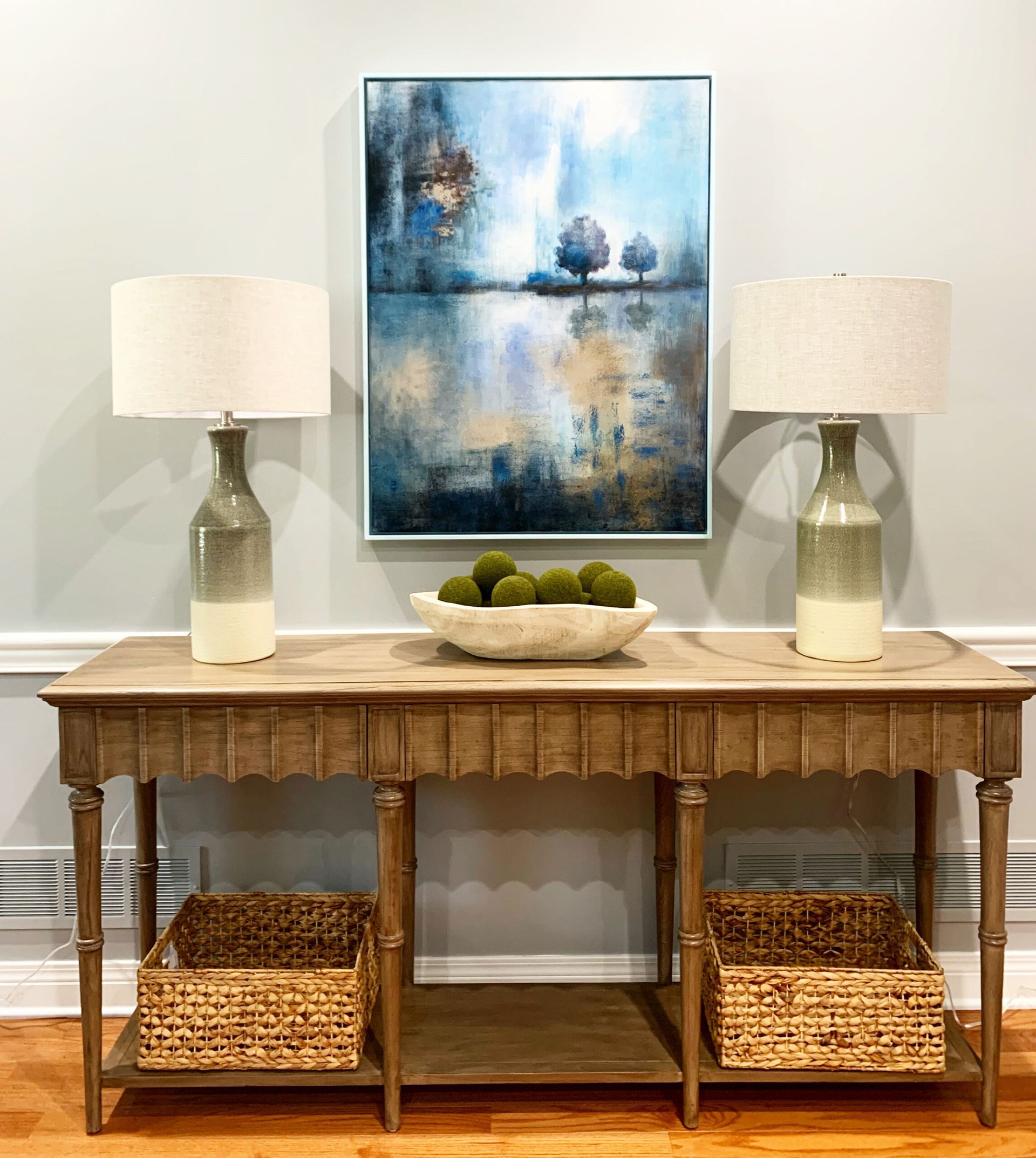 Console table with lamps, a blue abstract painting, and baskets in a bright entryway.