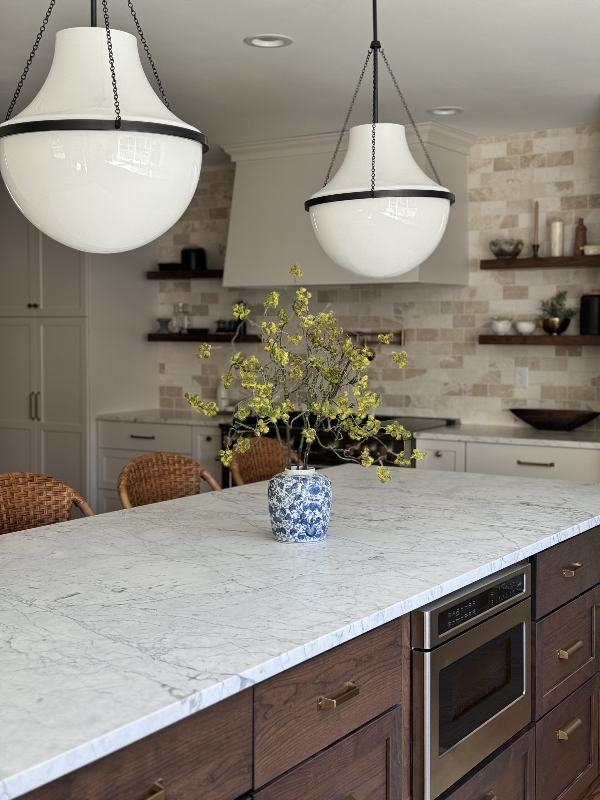 Bright kitchen with white marble island, pendant lights, open shelves, and a vase of yellow flowers.