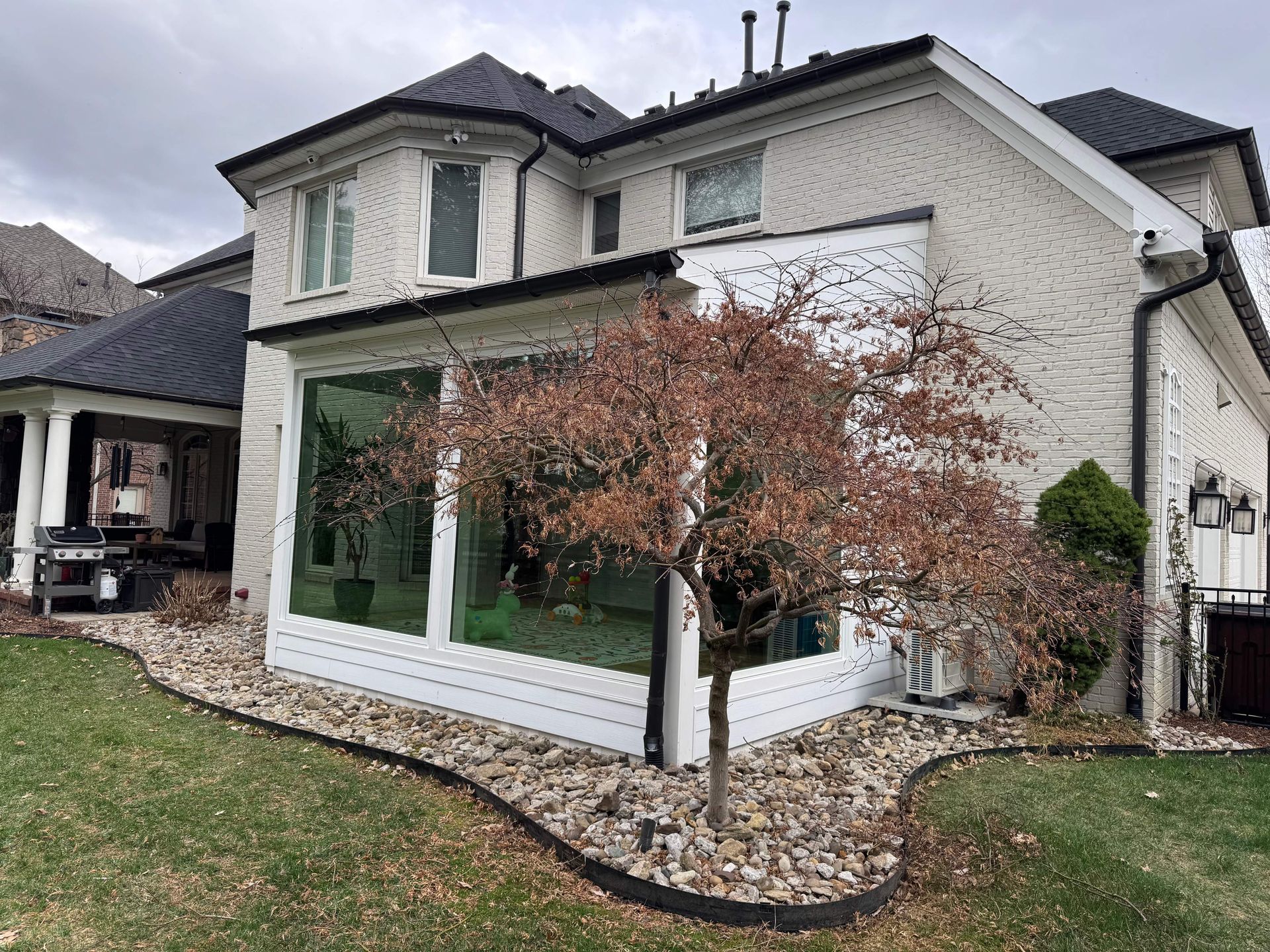 White two-story house with dark roof, large windows, and a small landscaped yard with a tree and rocks