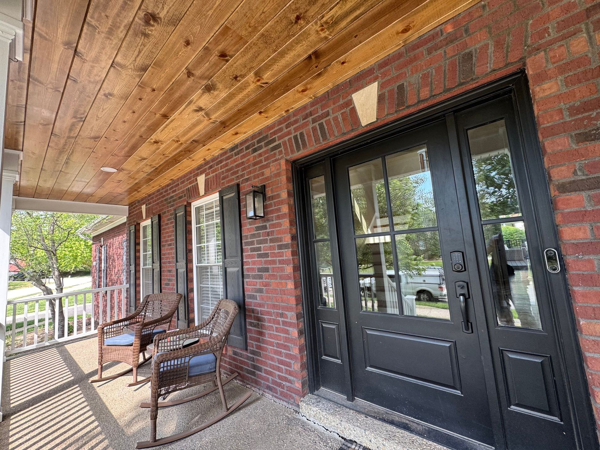 Covered brick porch with black double doors, wooden ceiling, and wicker chairs by a white railing.