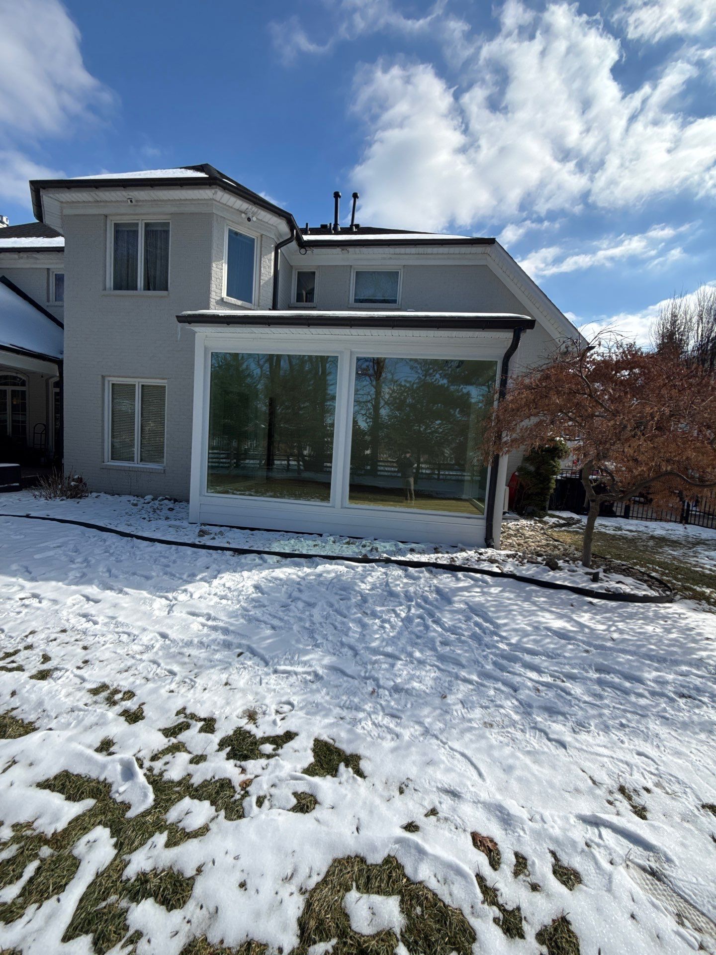 Snow-covered modern house with large glass windows and a red-leafed tree in front.
