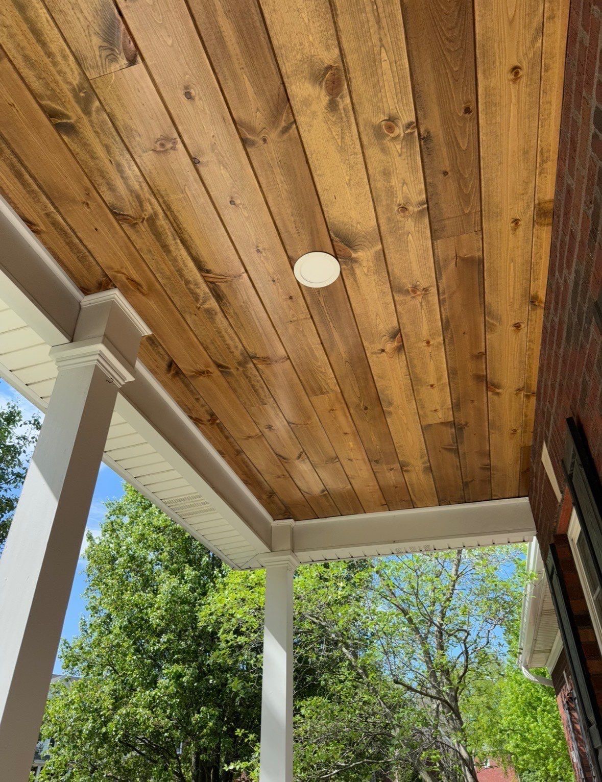 Wooden porch ceiling with a round light fixture, framed by white columns and green trees beyond.