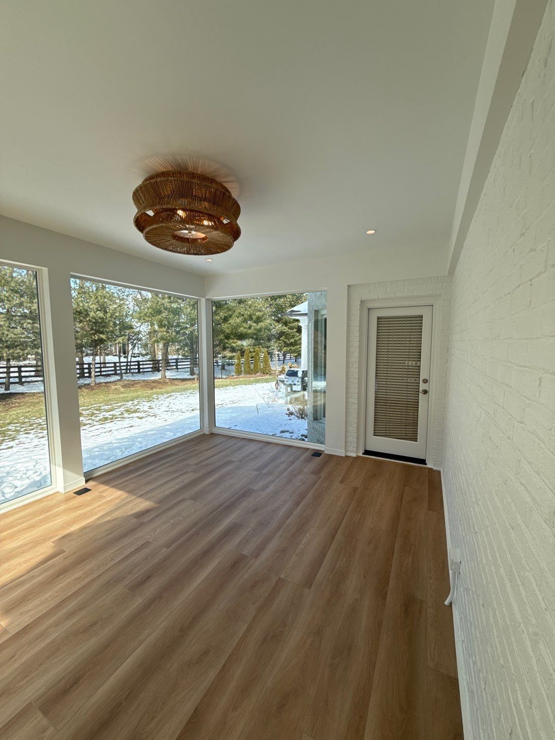 Empty sunroom with wood floors, wicker ceiling light, and large windows overlooking a snowy yard
