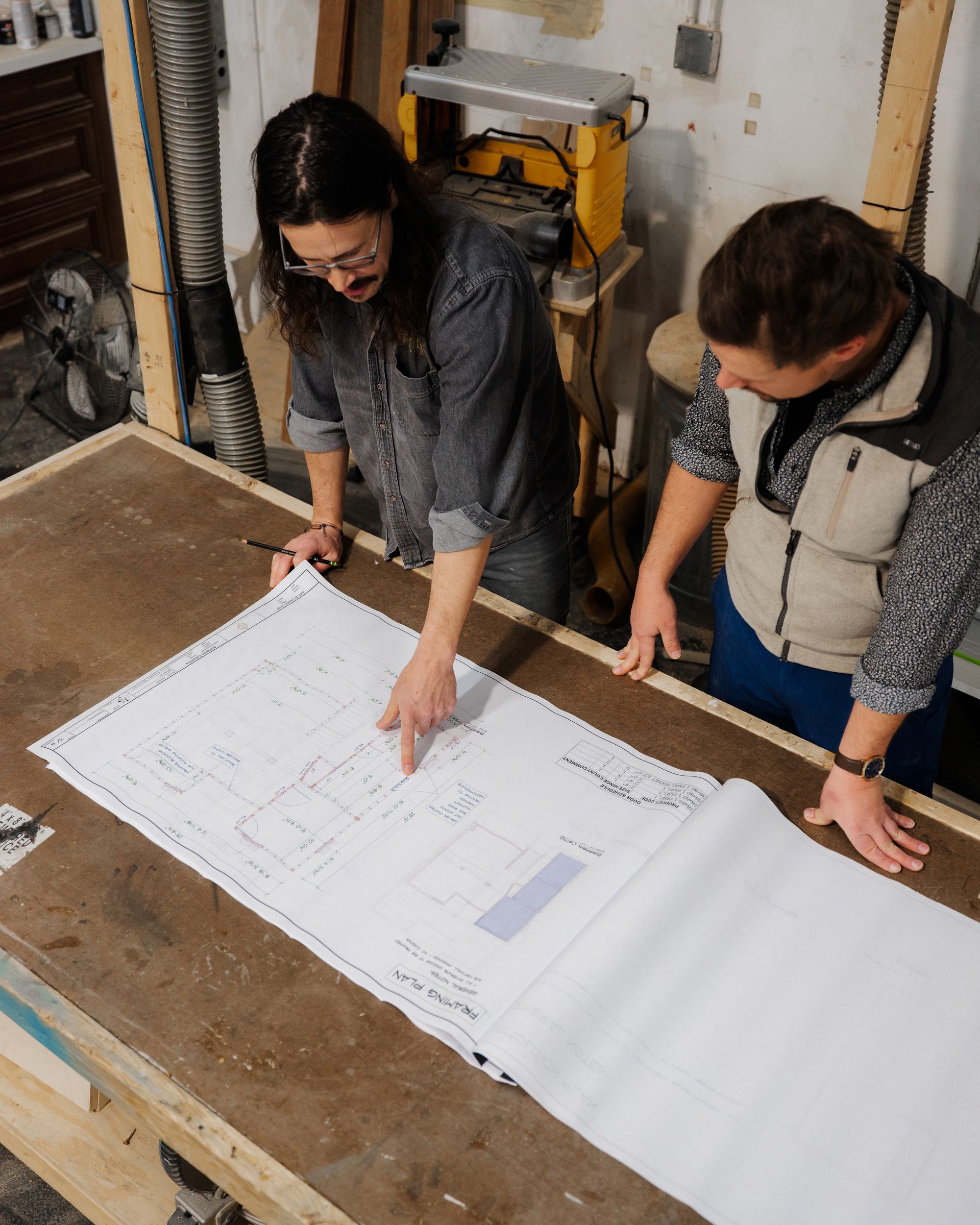 Two people reviewing a large blueprint spread across a workbench in a workshop.