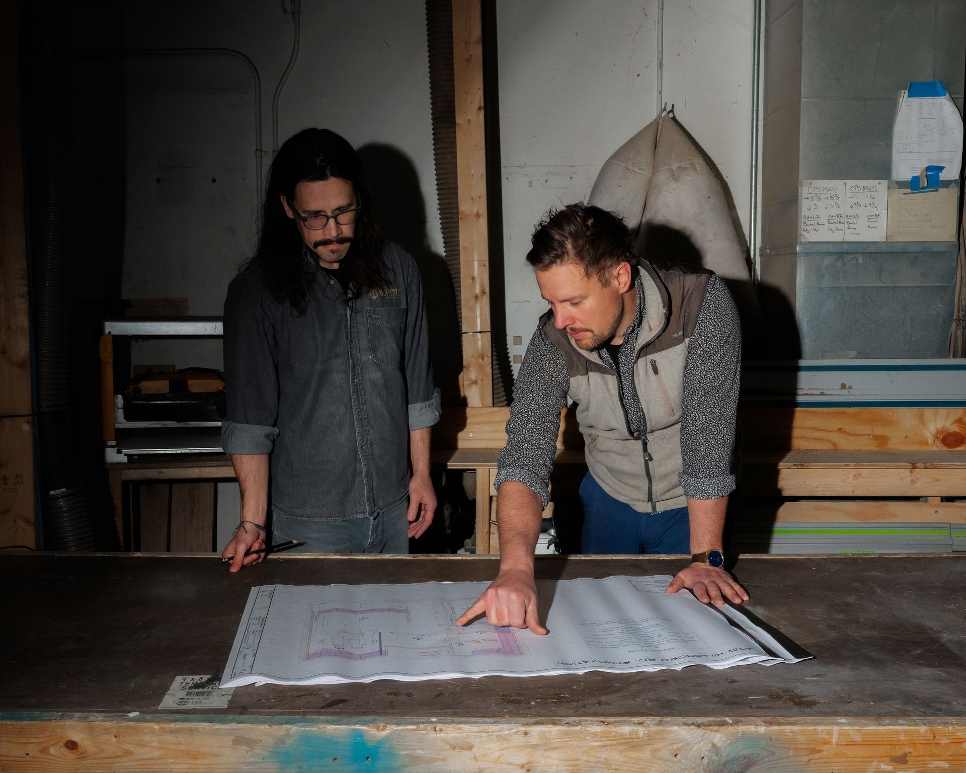 Two people lean over a large blueprint on a worktable in a dim workshop