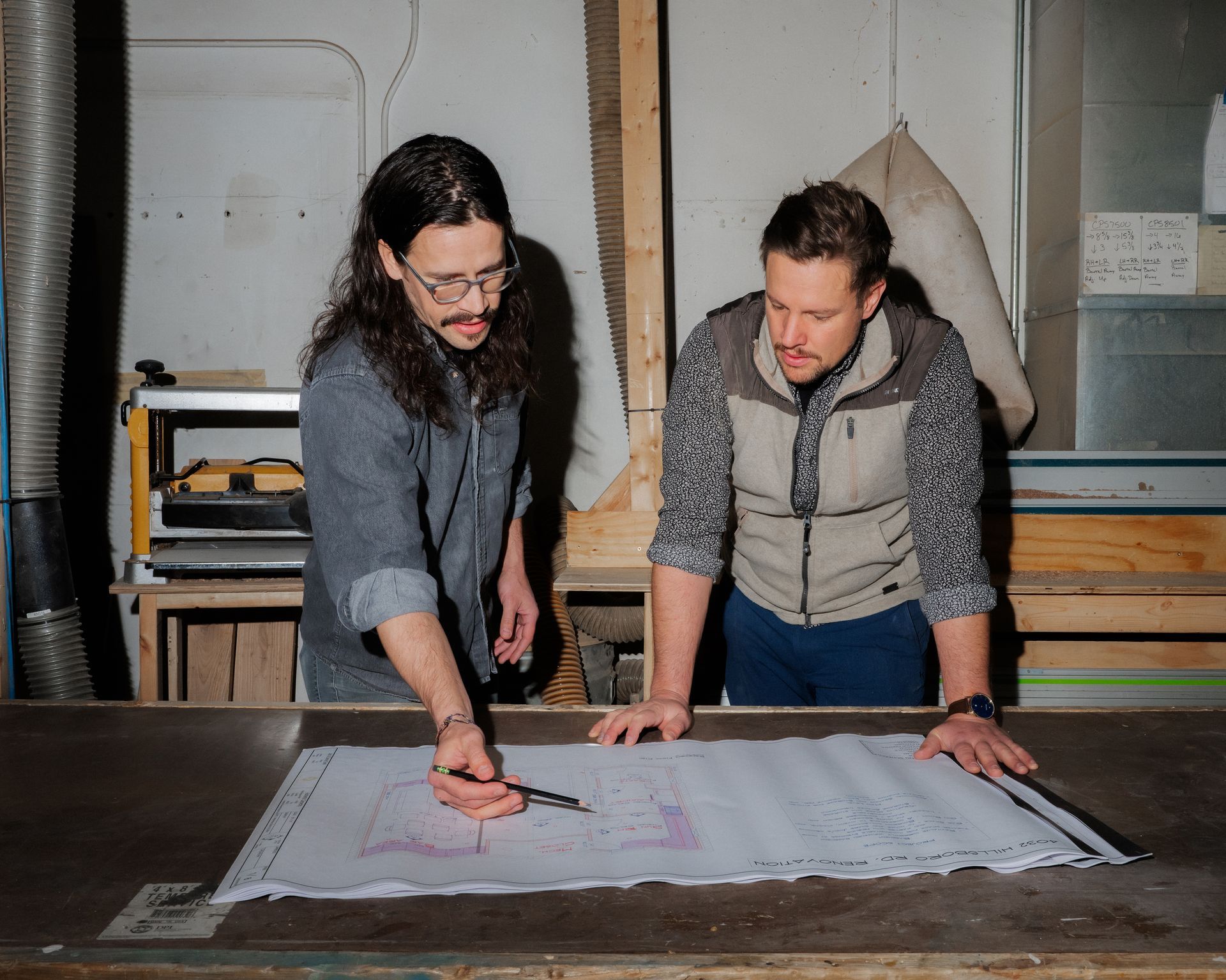 Two people leaning over a large floor plan, discussing and pointing at details in a workshop.