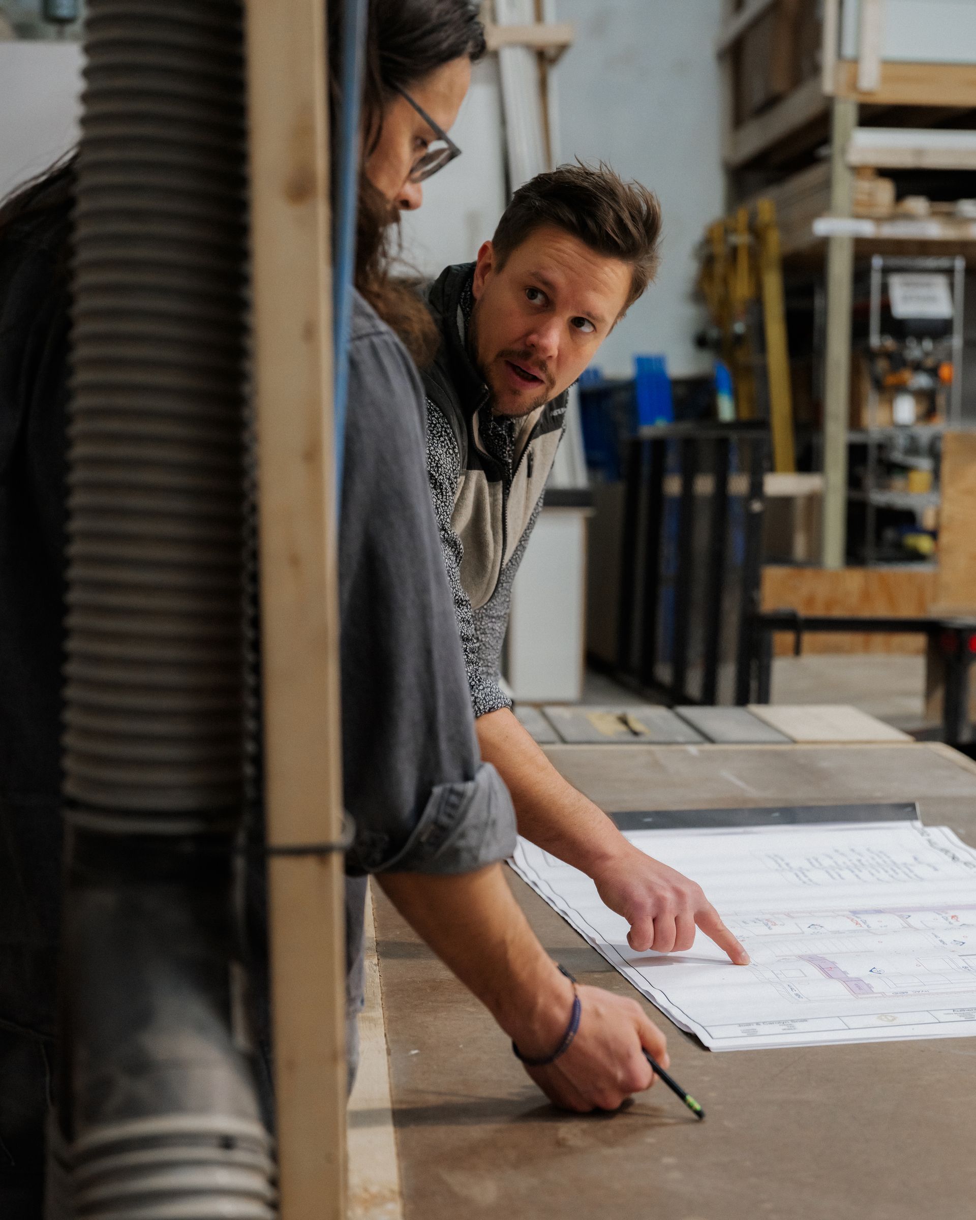 Two coworkers review blueprints on a table in a workshop, one pointing while the other writes.