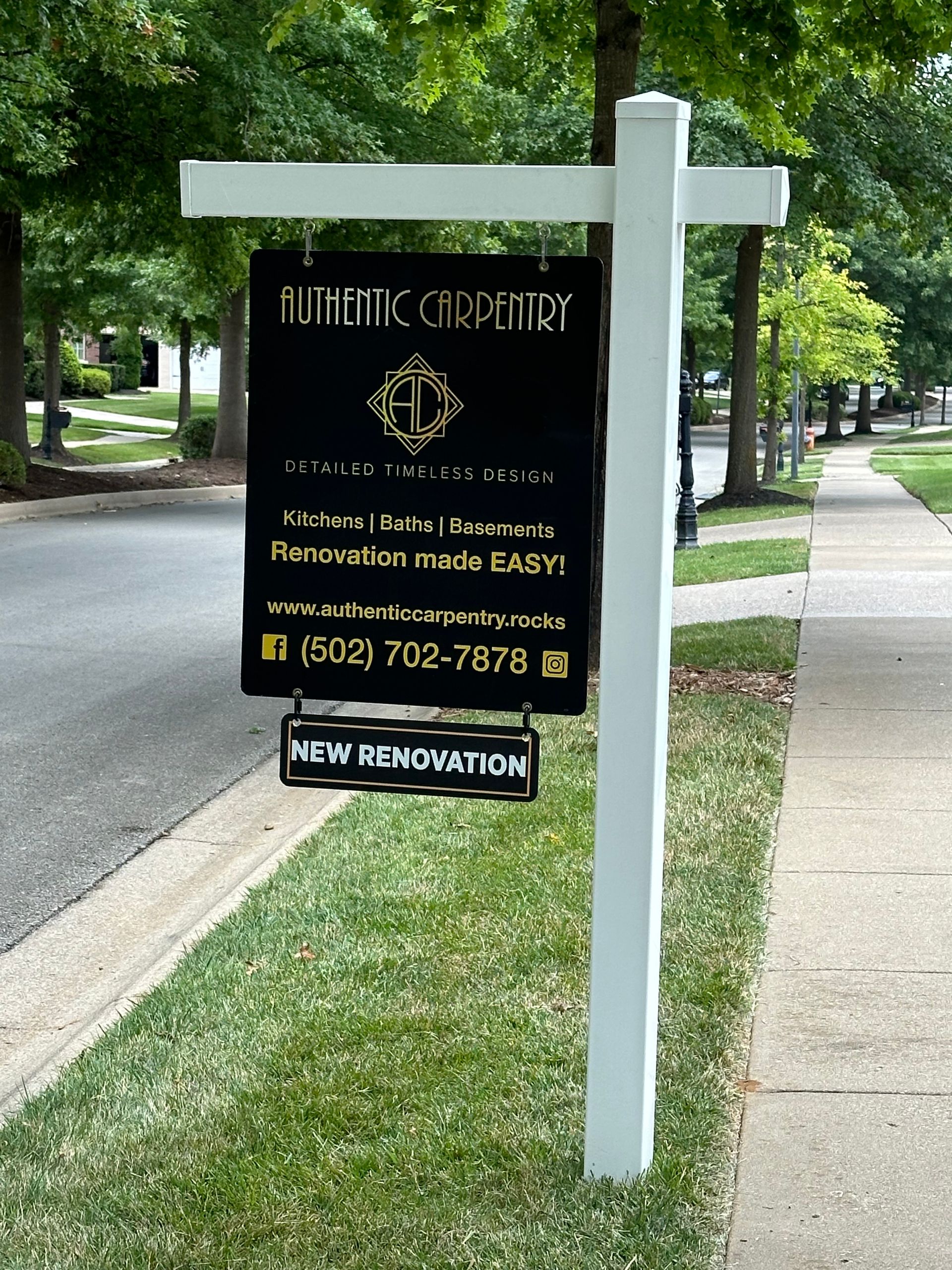 Black realty sign hanging from a white post on a tree-lined sidewalk.