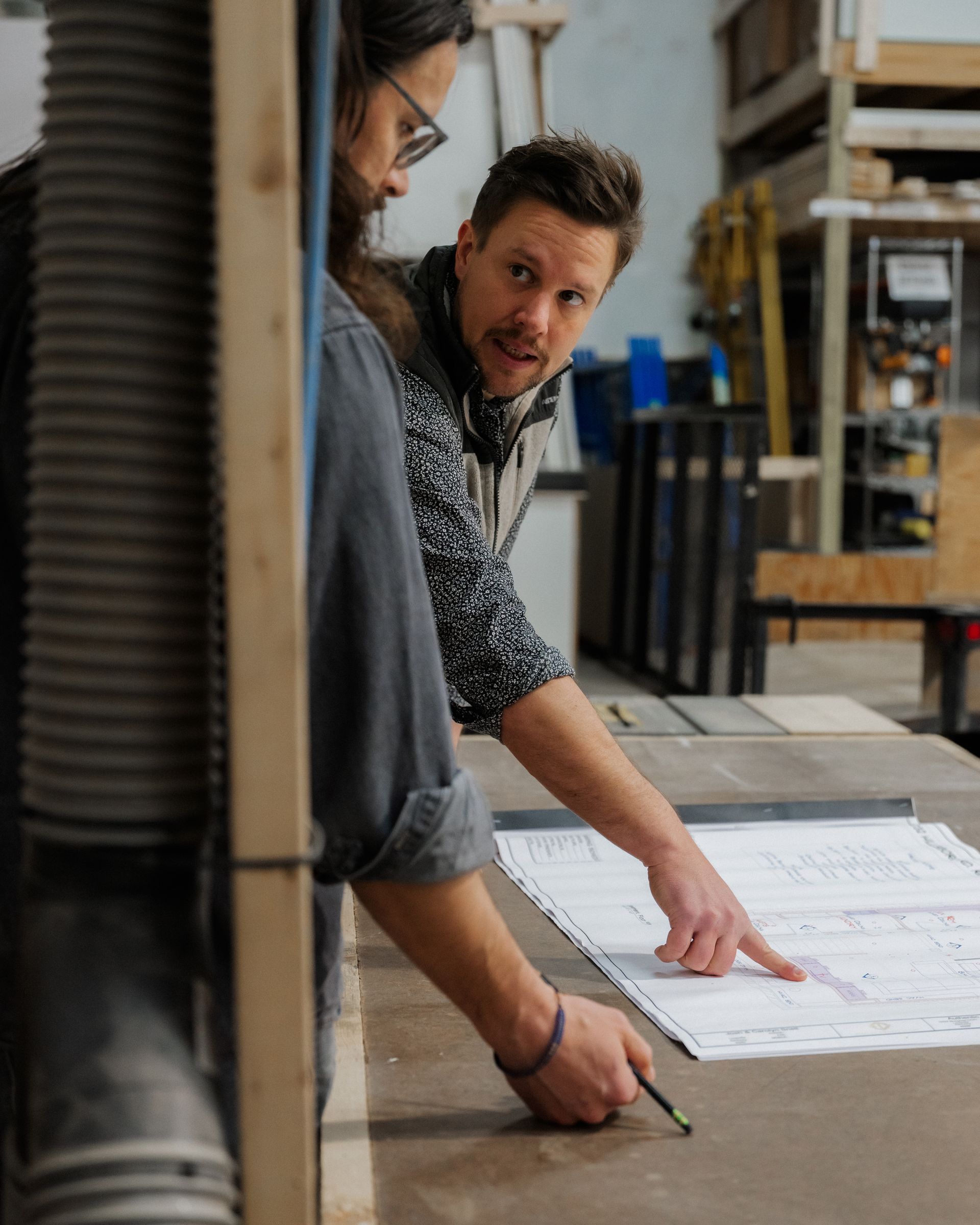 Two people at a workbench reviewing plans, one pointing at blueprints in a workshop