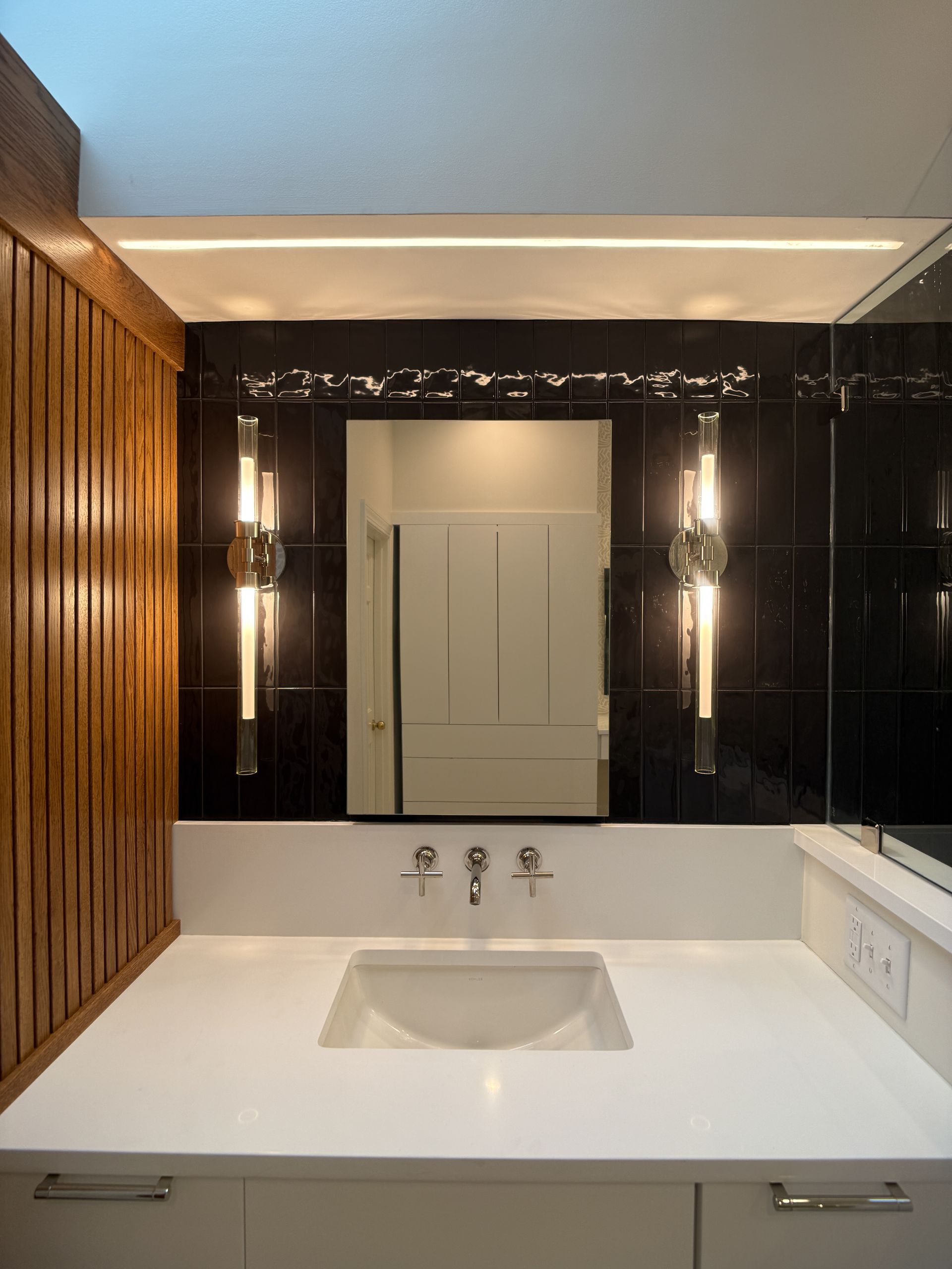 Modern bathroom vanity with white sink, black mirror wall, and vertical sconces beneath a lit ceiling niche