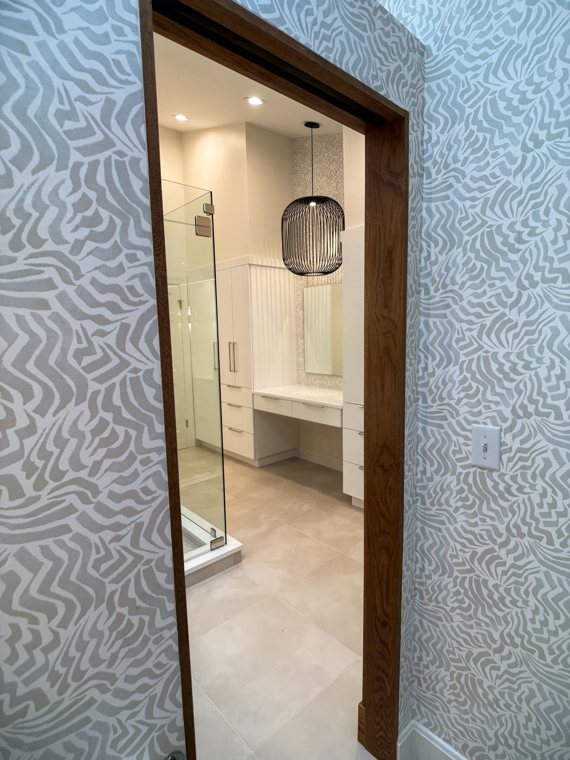 Bathroom vanity seen through a wooden doorway, with glass shower and patterned wallpaper visible.