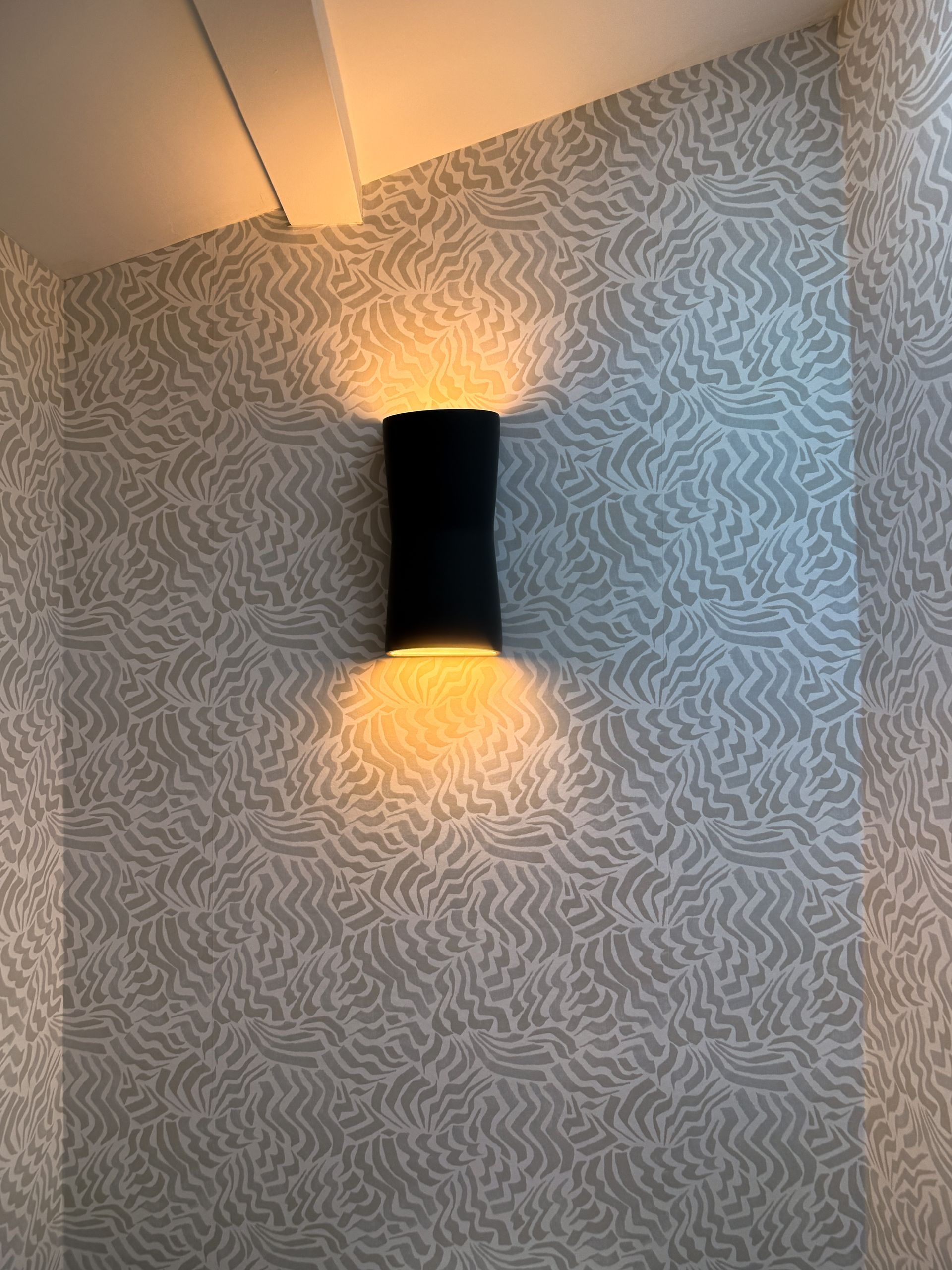 Black wall sconce glowing on textured gray wallpaper, casting warm light