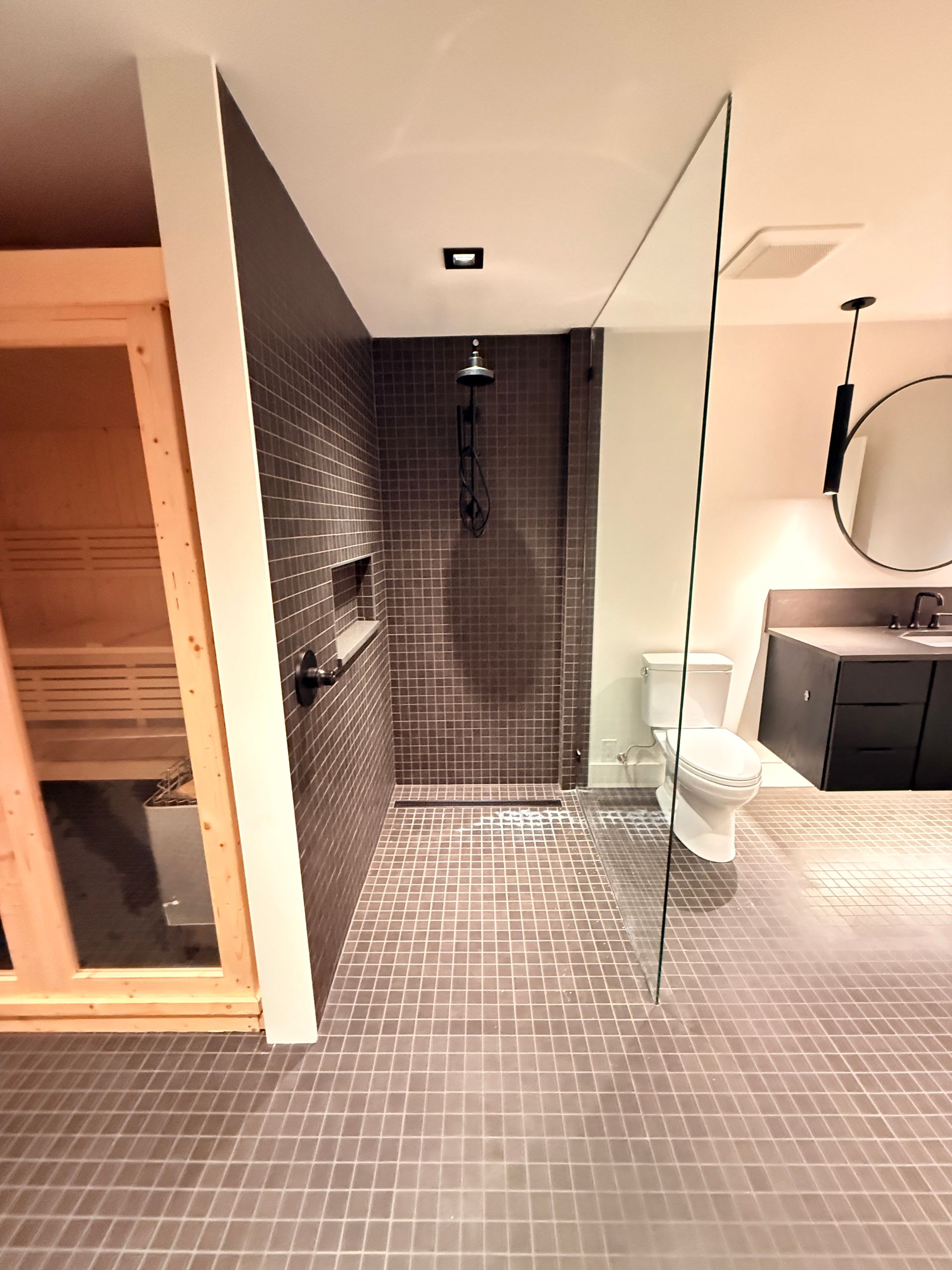Modern bathroom with black tile shower, glass partition, toilet, vanity, and wooden sauna area