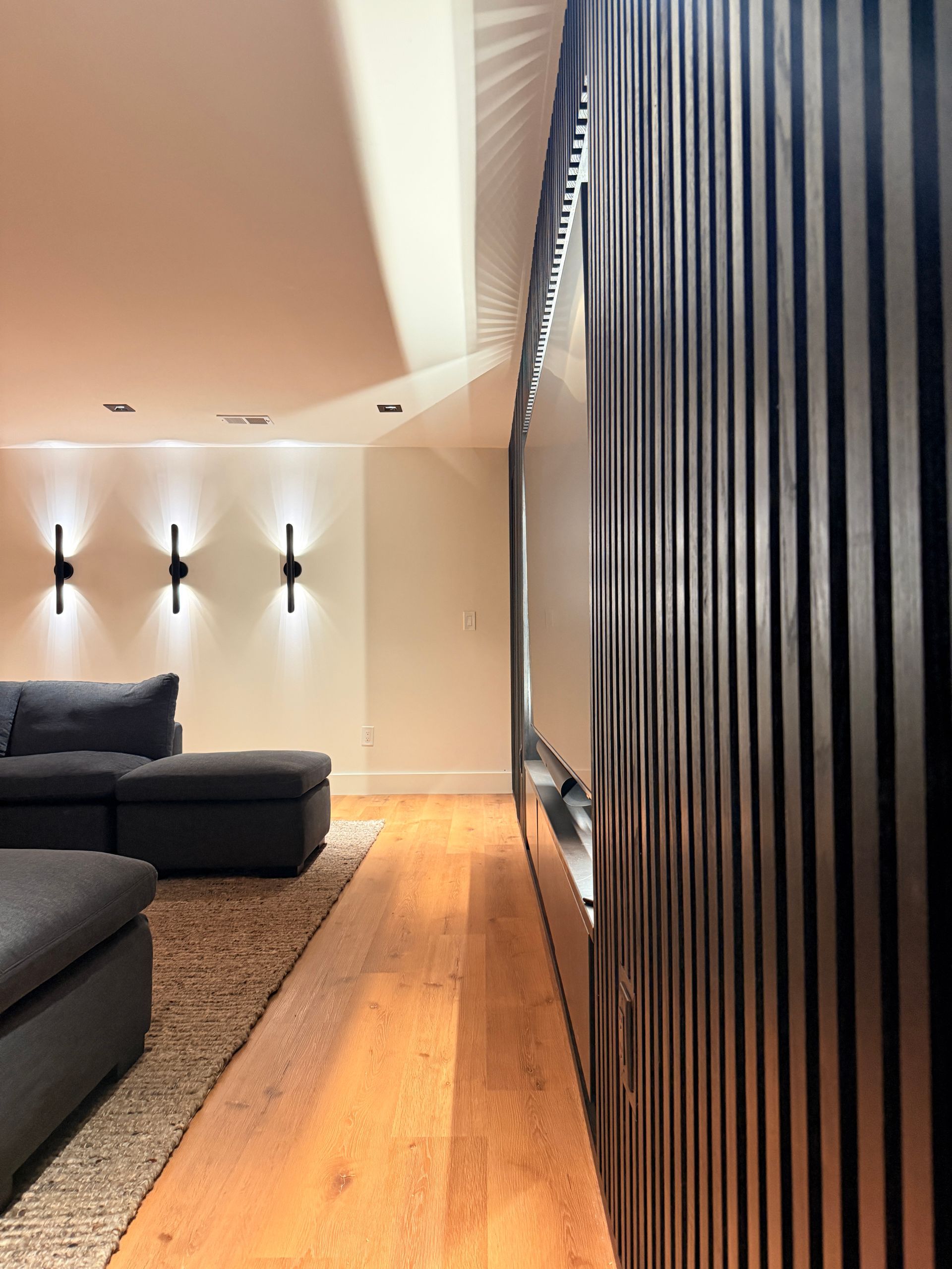 Modern living room with dark sectional sofa, wall sconces, wooden floor, and black slatted partition