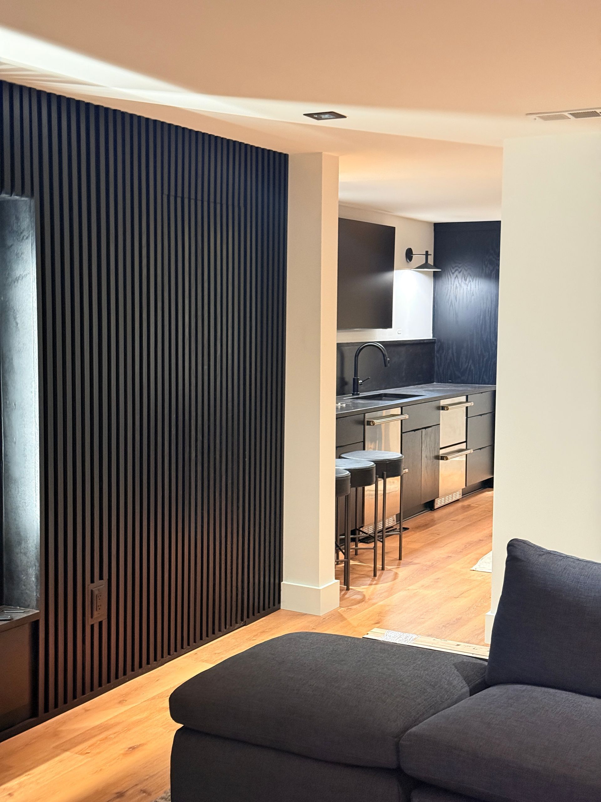 Modern living room with dark sectional, wood floor, vertical blinds, and a view into a compact kitchen.