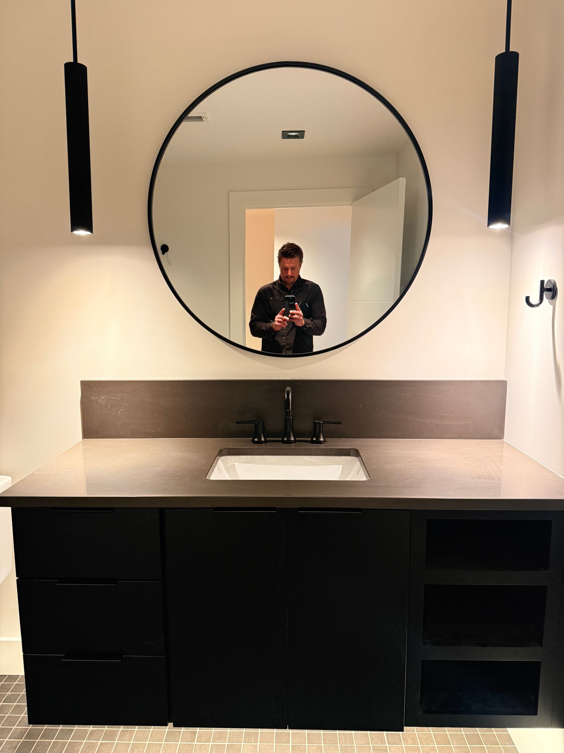 Modern bathroom vanity with round mirror, black fixtures, and hanging pendant lights, reflecting a person taking a photo