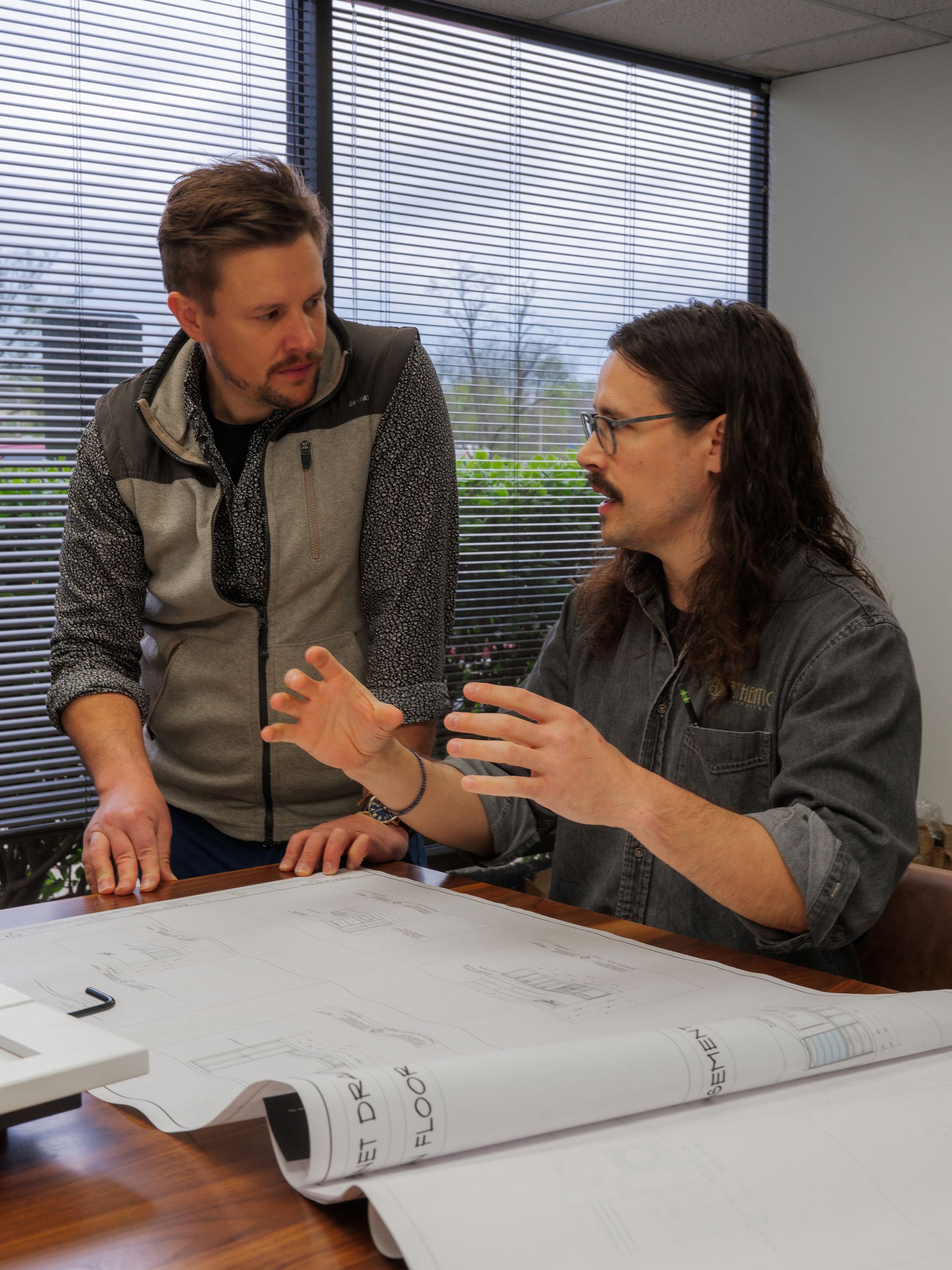 Two coworkers discuss plans at a table with large papers spread out in an office.
