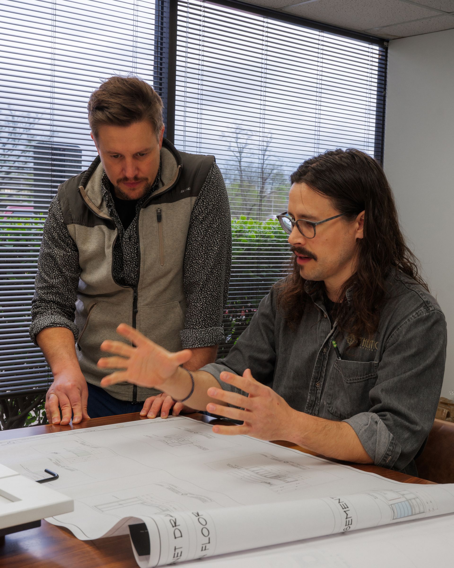 Two people reviewing large architectural plans at a desk in a bright office meeting room