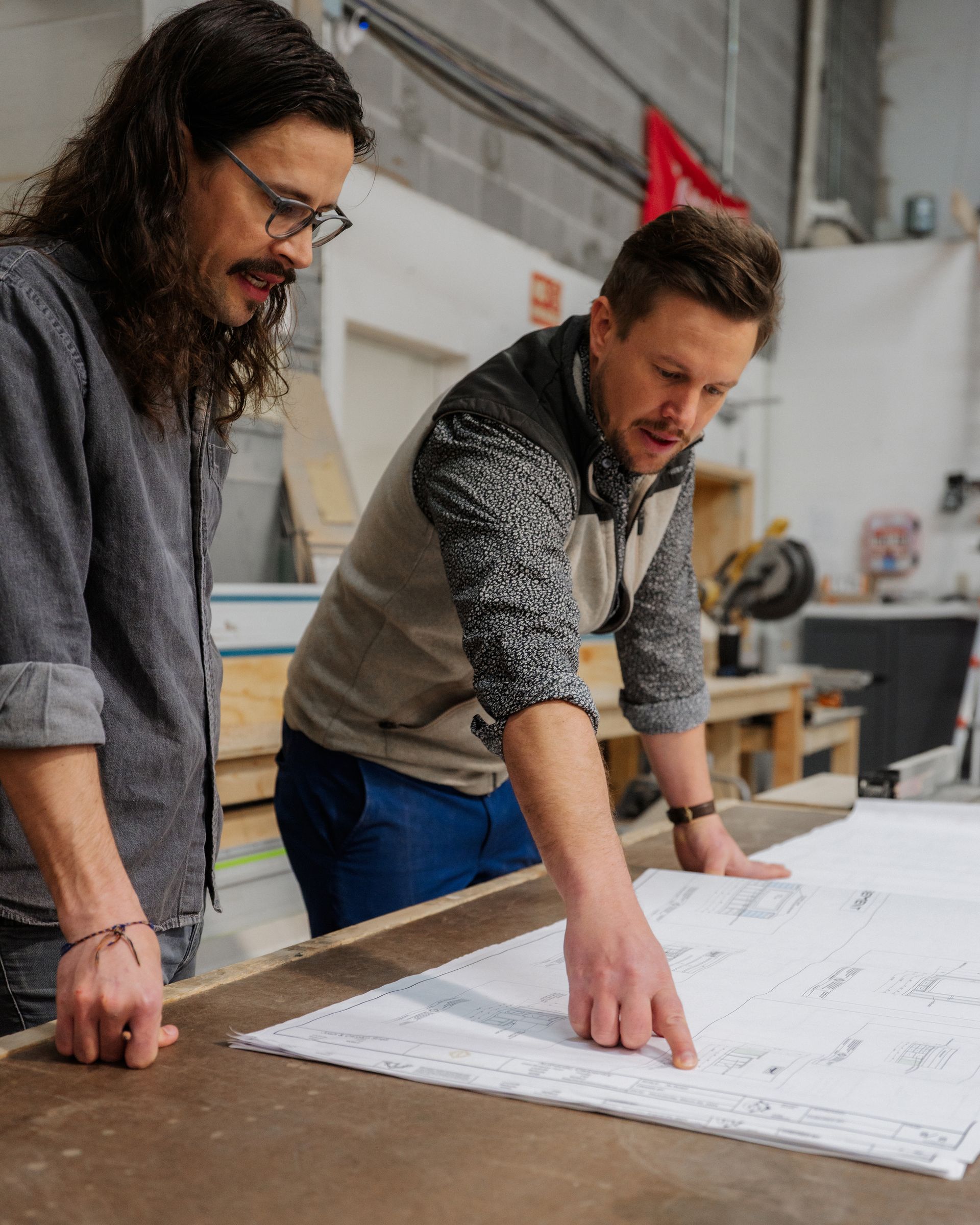 Two people leaning over a large blueprint in a workshop, pointing and discussing plans.