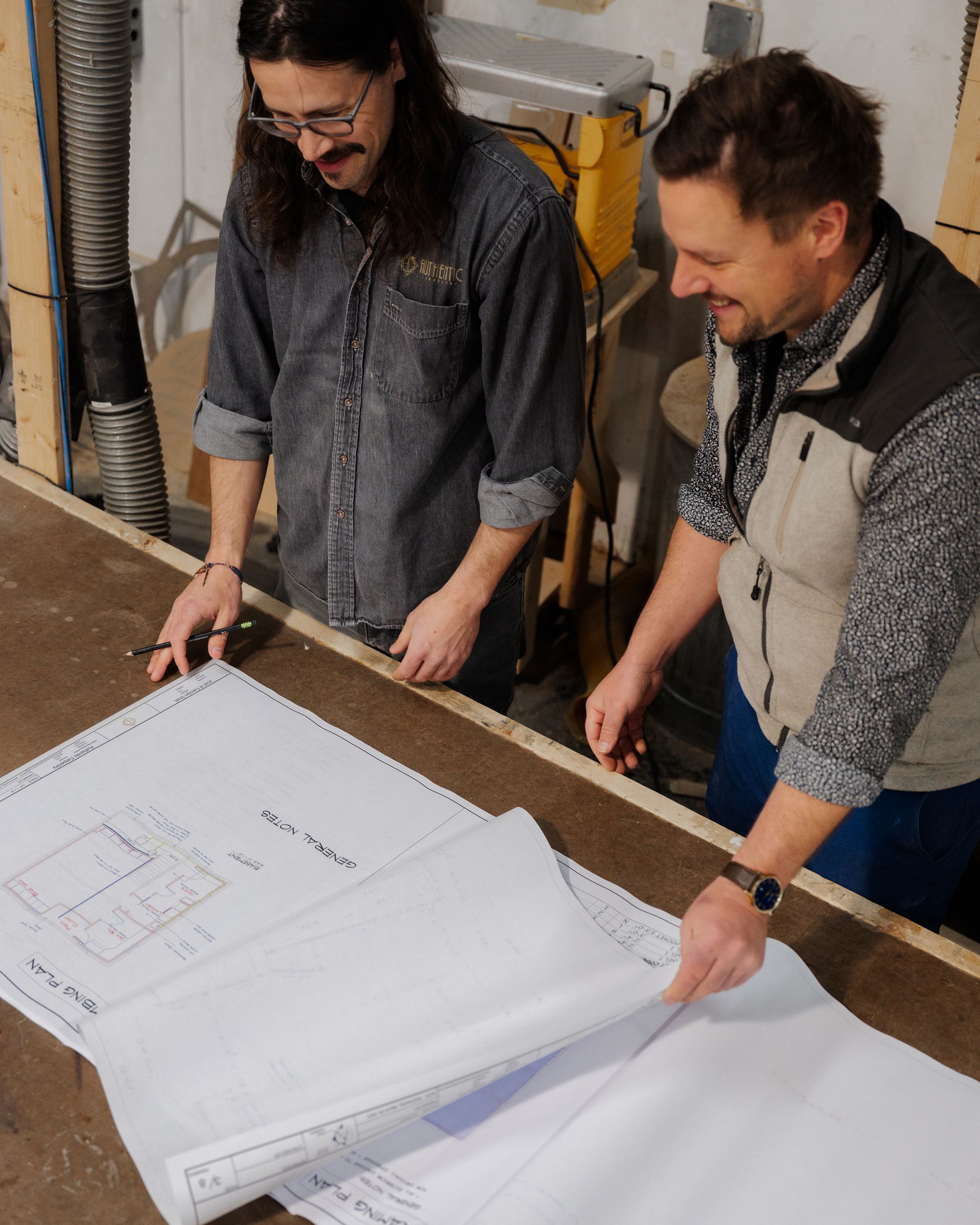 Two people reviewing large blueprints on a table in a workshop
