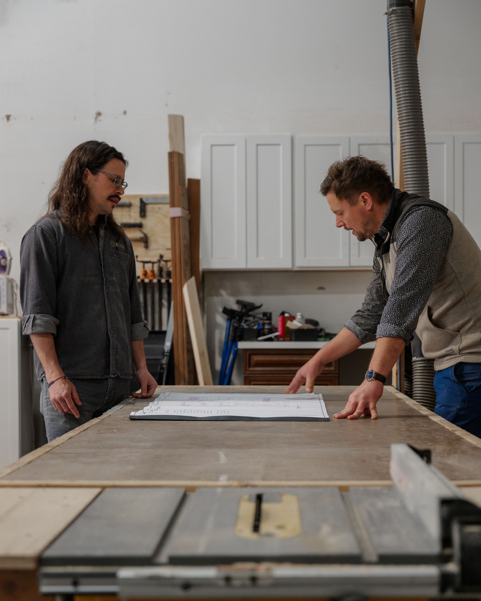 Two people measuring wood on a workbench in a workshop with tools and cabinets.