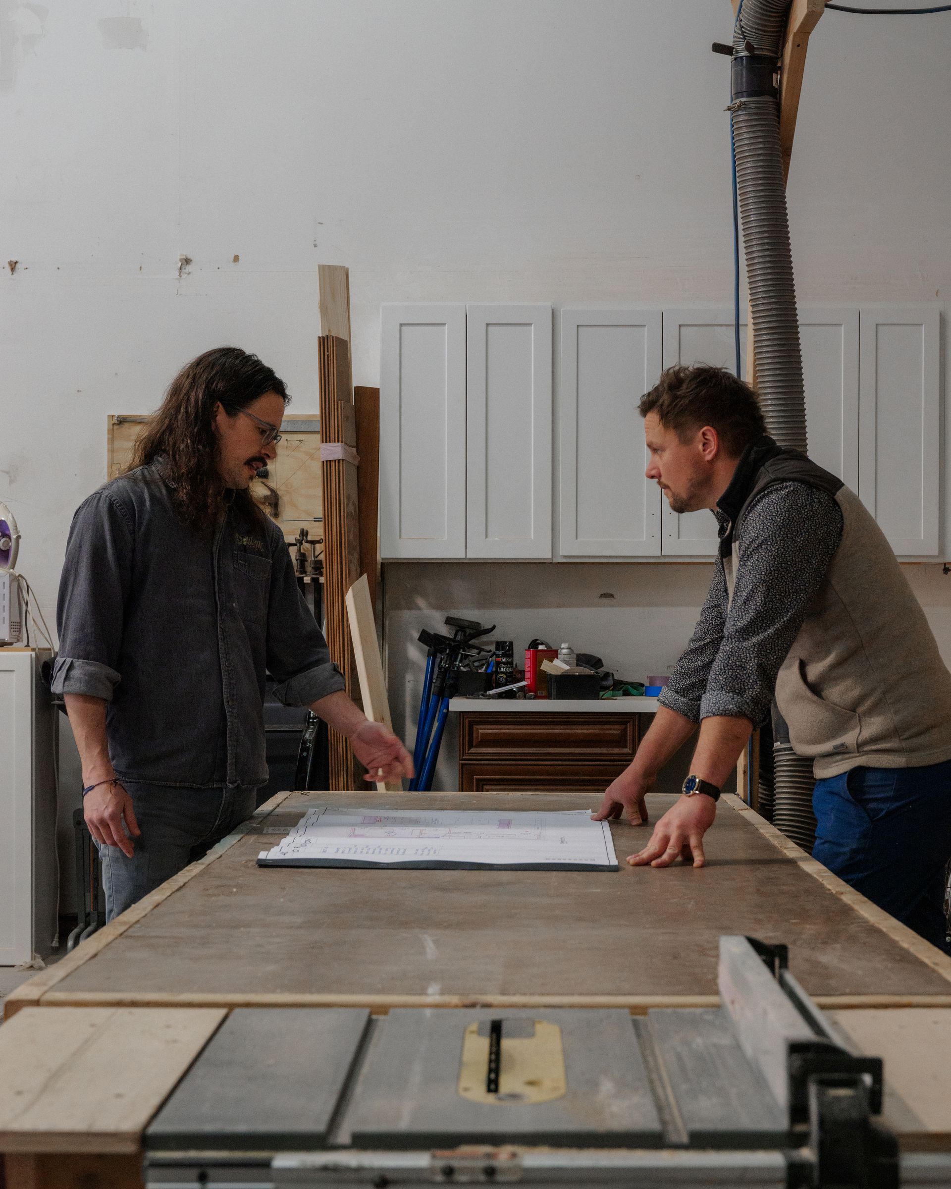 Two people in a workshop lean over a worktable with a large plan or board.