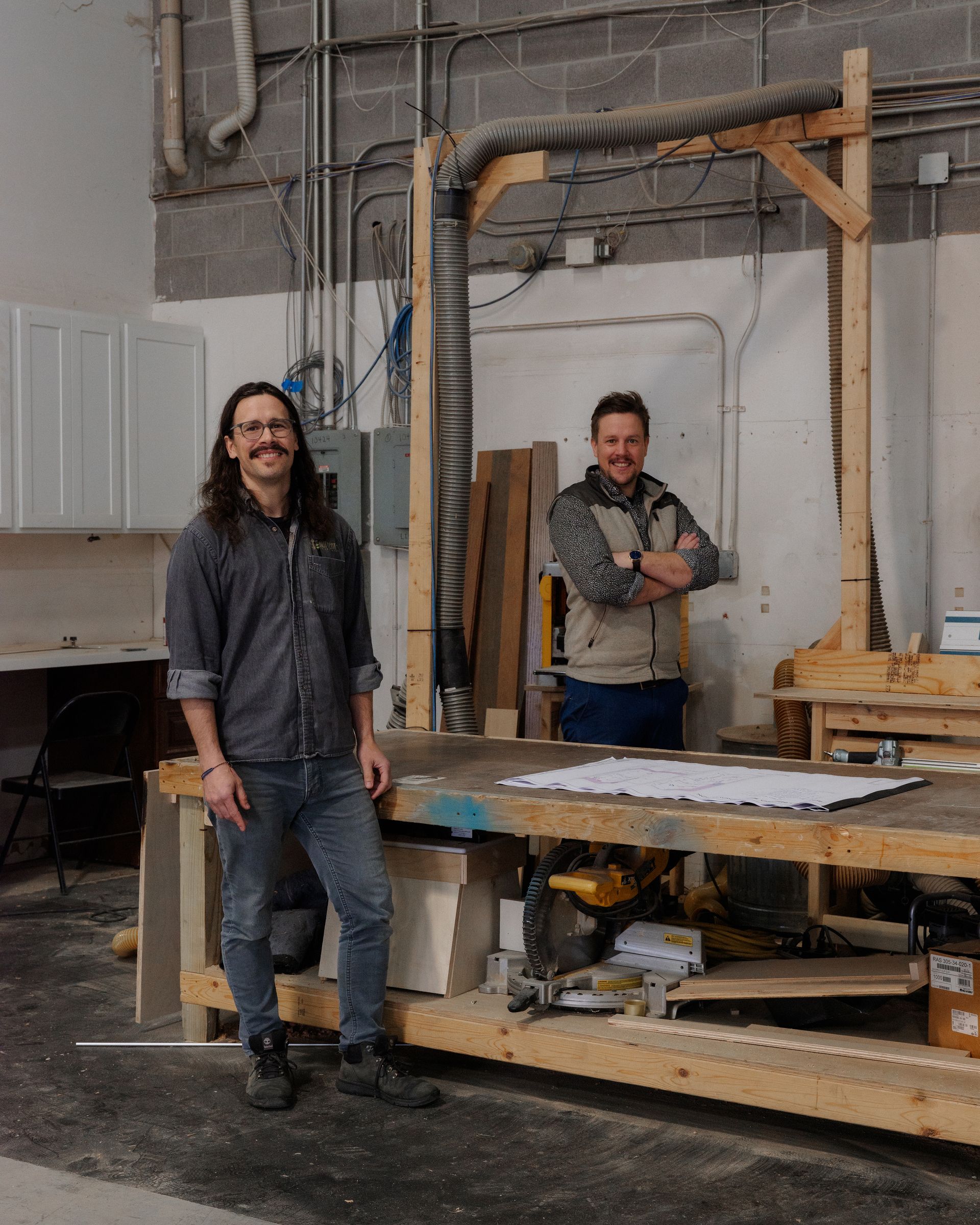 Two people stand in a workshop beside a wooden worktable and tools.
