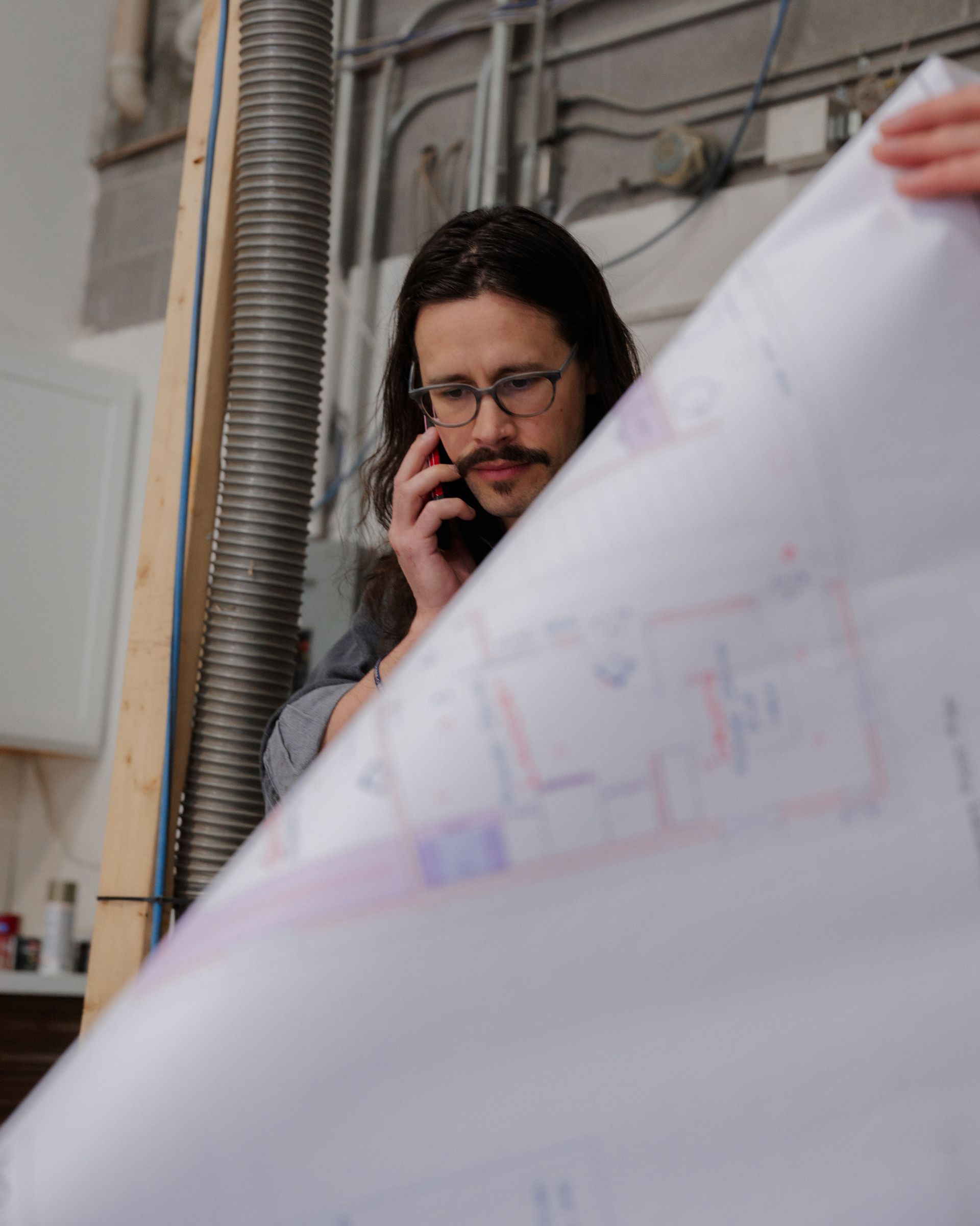 Person on a phone call reviewing a large blueprint in an industrial workspace.