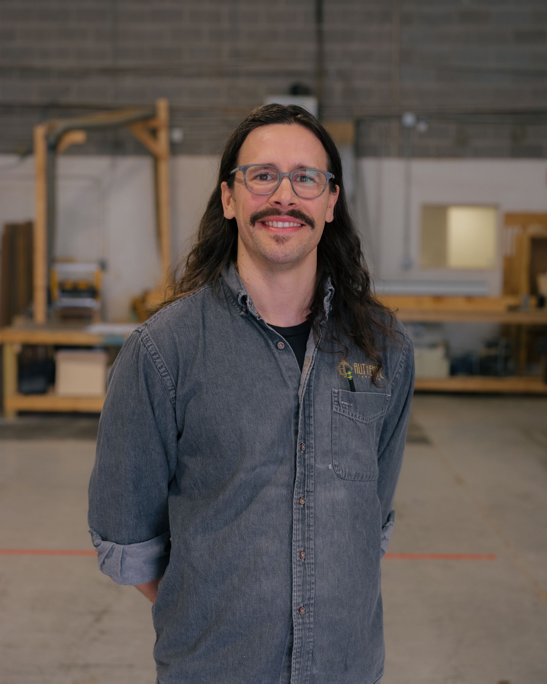 Smiling person with long dark hair and glasses standing in a workshop, wearing a gray shirt.