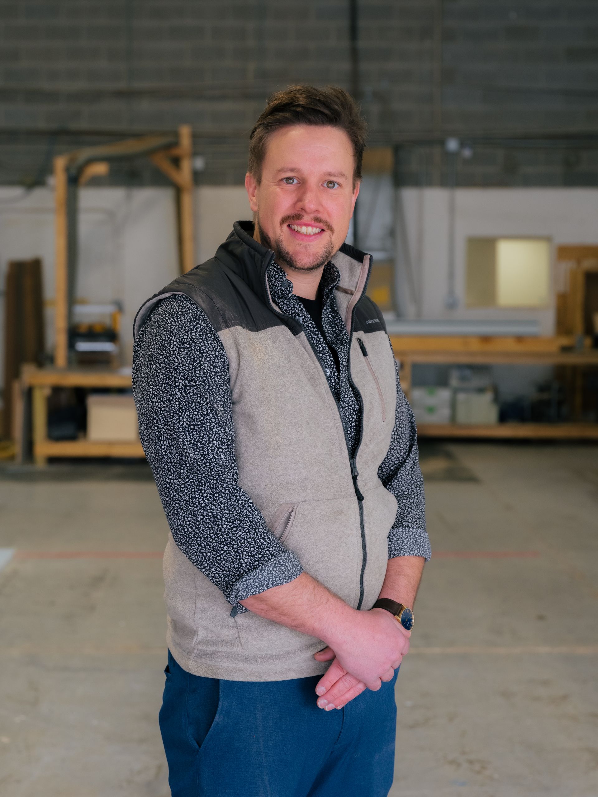 Smiling man in a gray vest standing in an industrial workshop with wooden shelving in the background