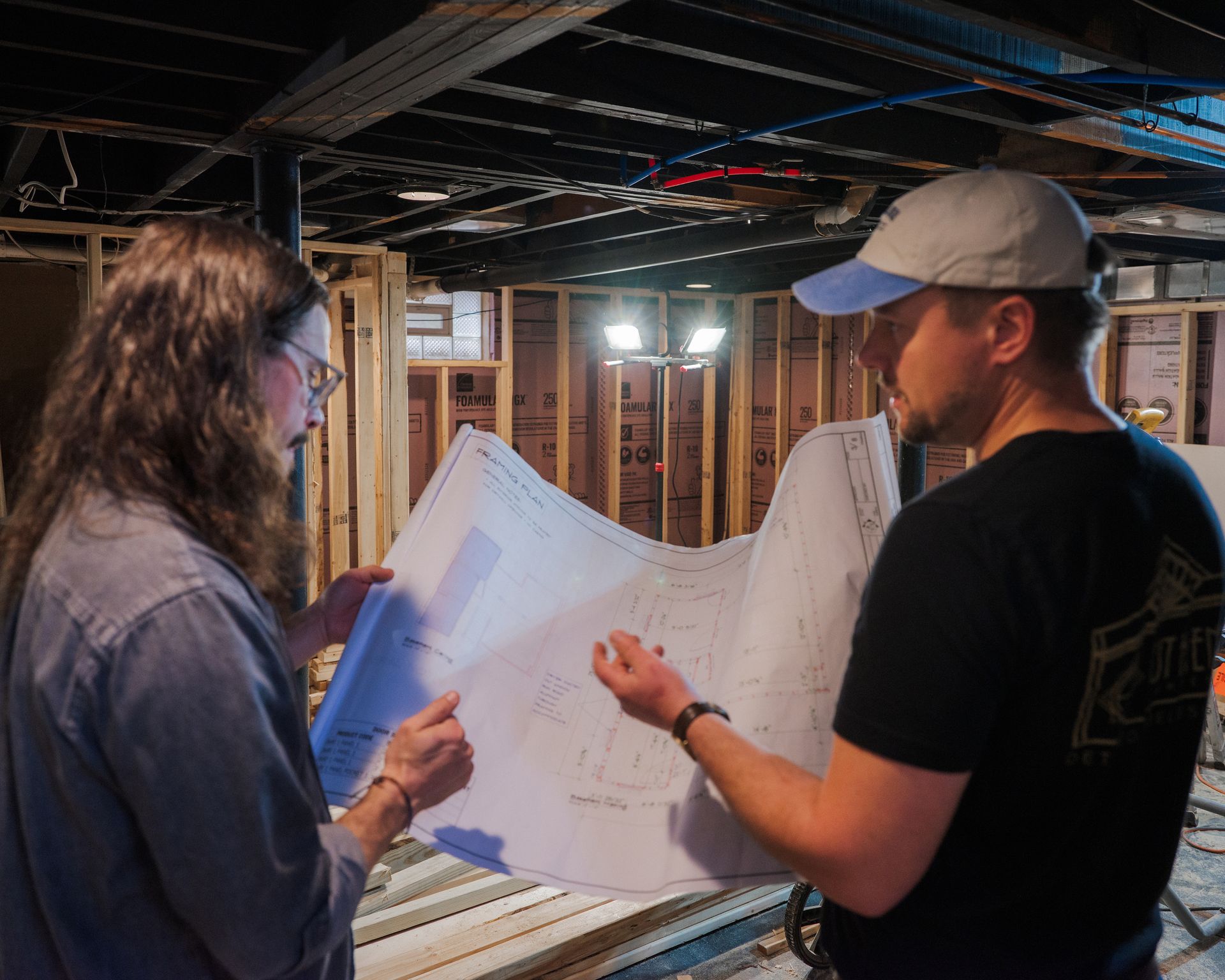 Two people reviewing large blueprints inside a construction site with exposed framing and ceiling pipes
