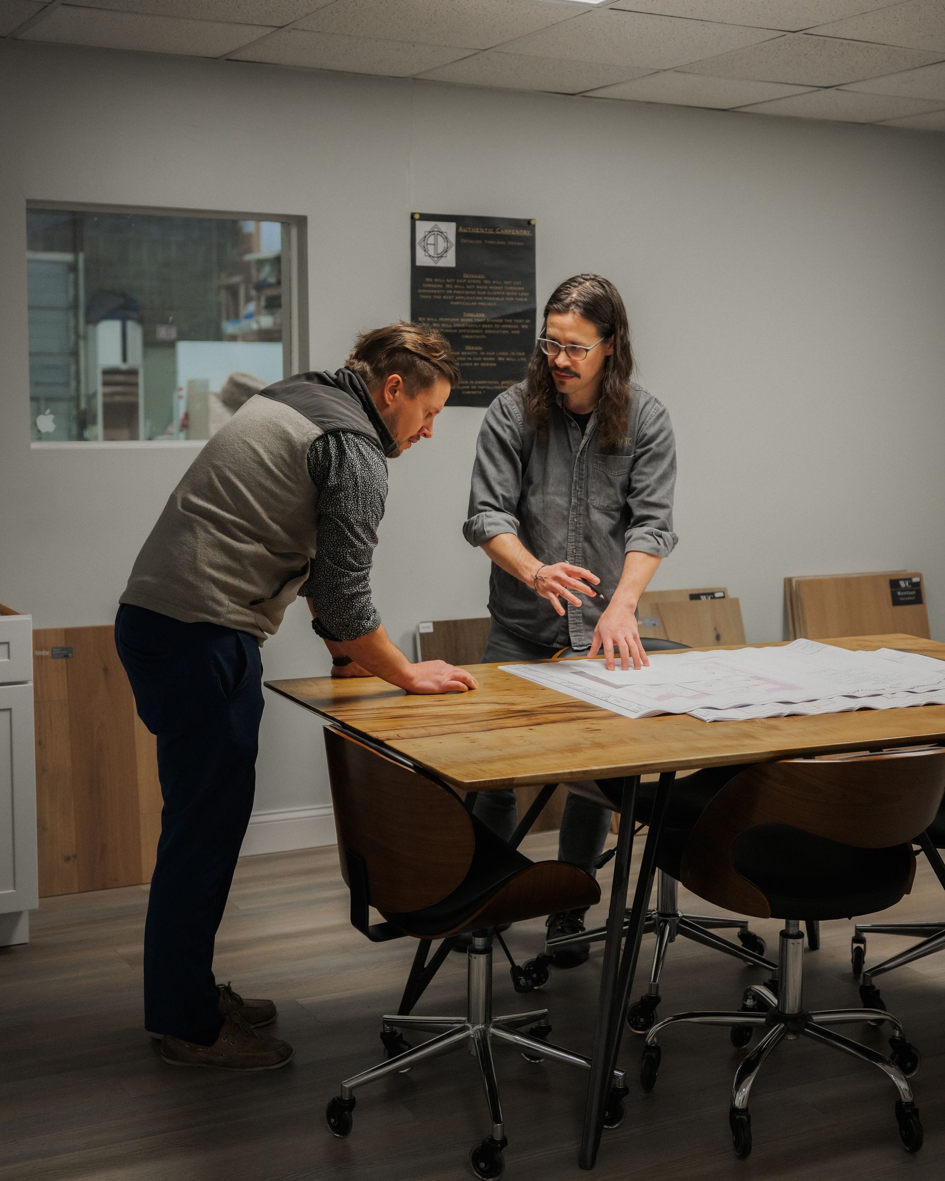 Two people lean over a large table, reviewing papers in a conference room.