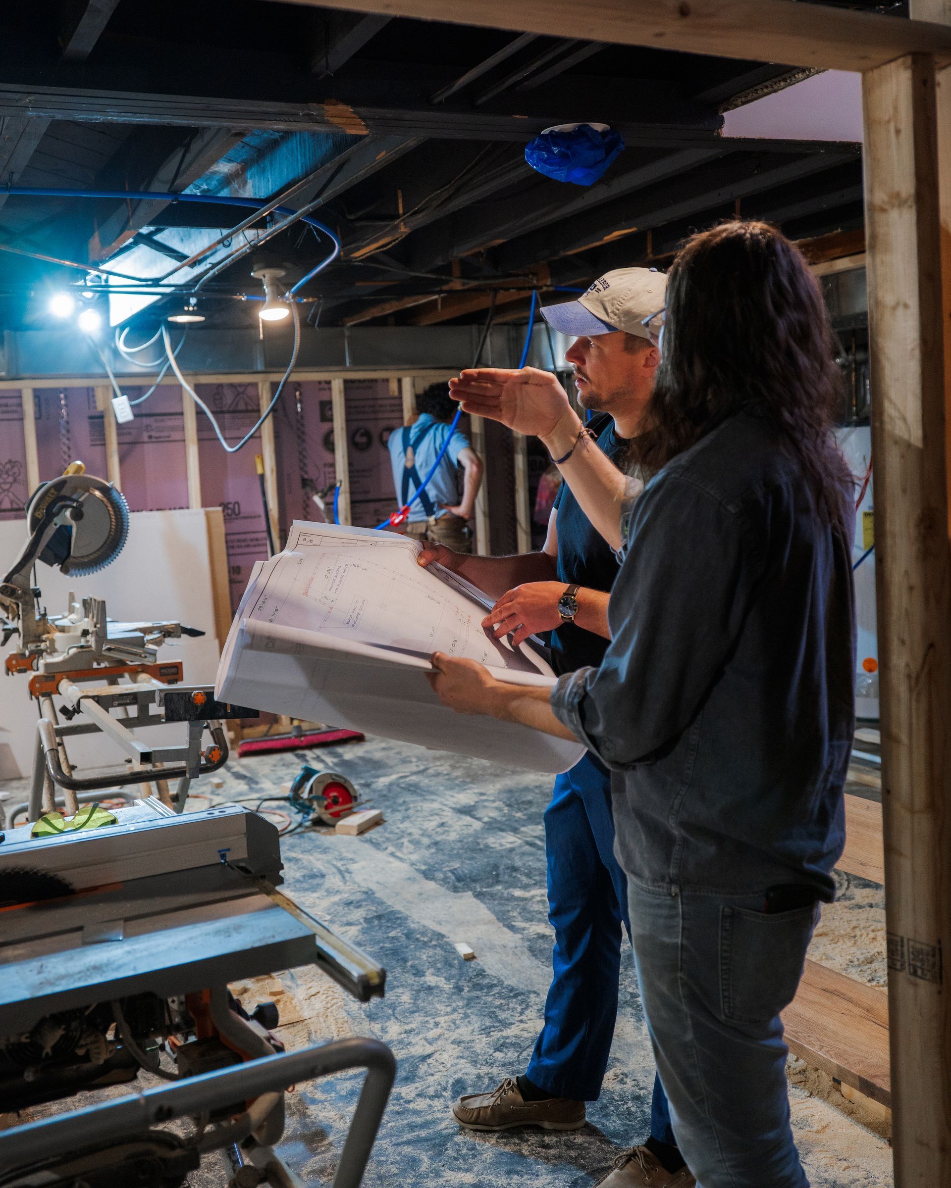 Two people reviewing a large plan in a cluttered workshop under exposed beams.