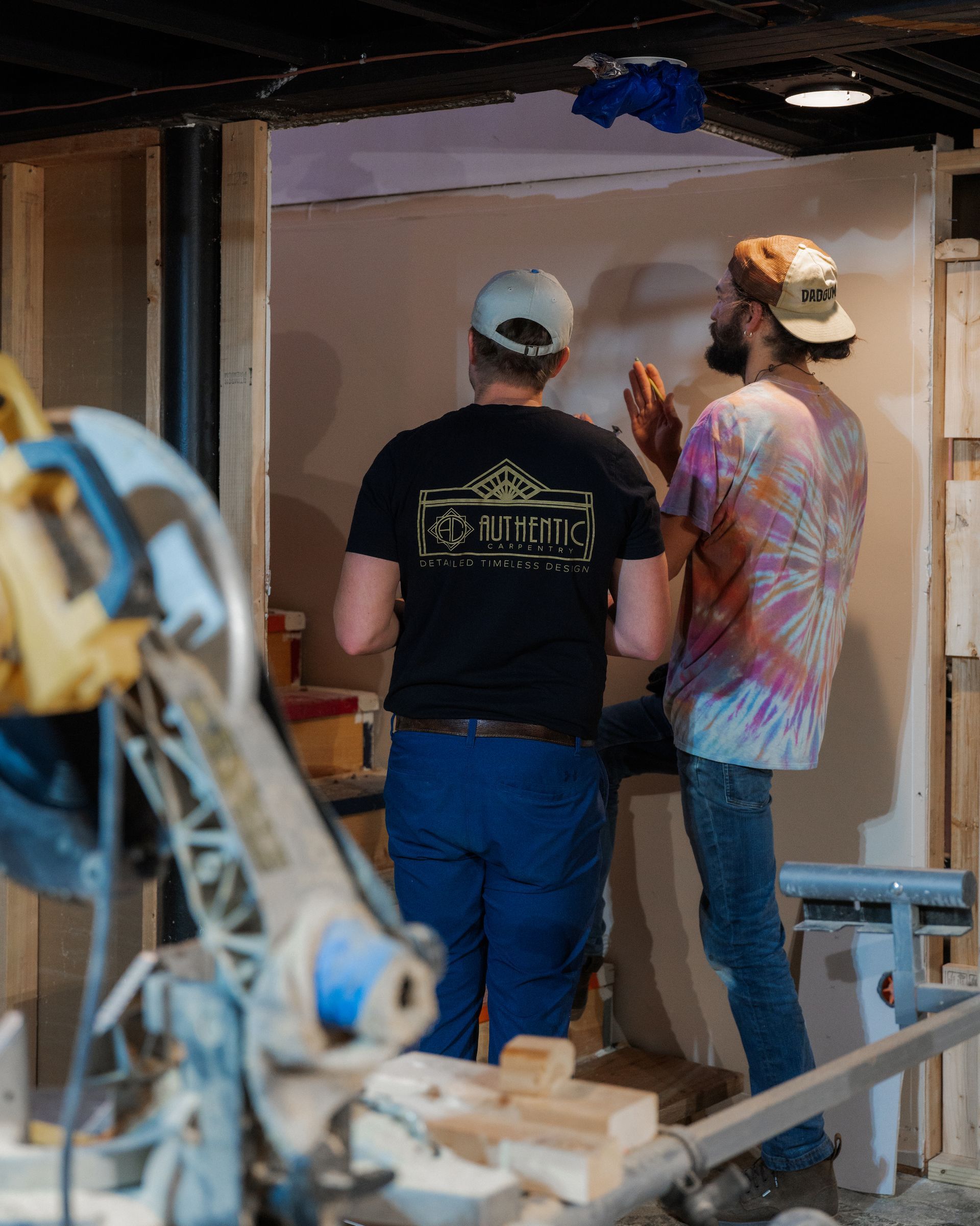 Two people in caps stand in a renovation site, examining a wall near scaffolding and tools.