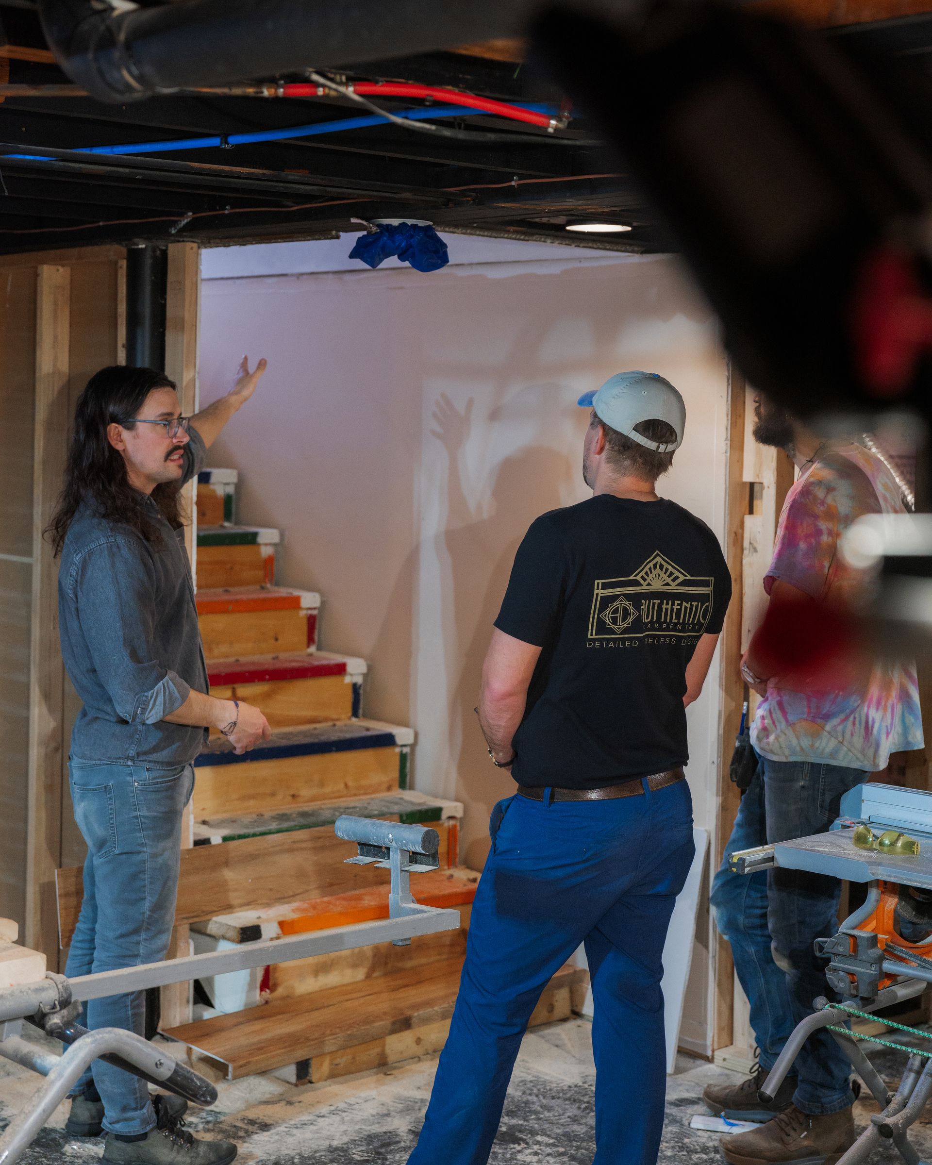 Two people examining a wall in a basement workshop, with wood steps and tools nearby.