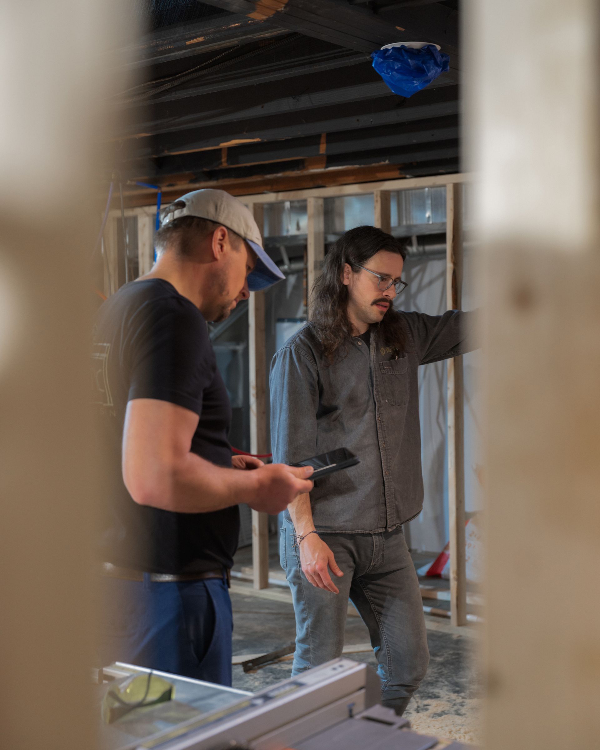 Two people in a construction site, one pointing at the unfinished wall while the other looks down at a phone.