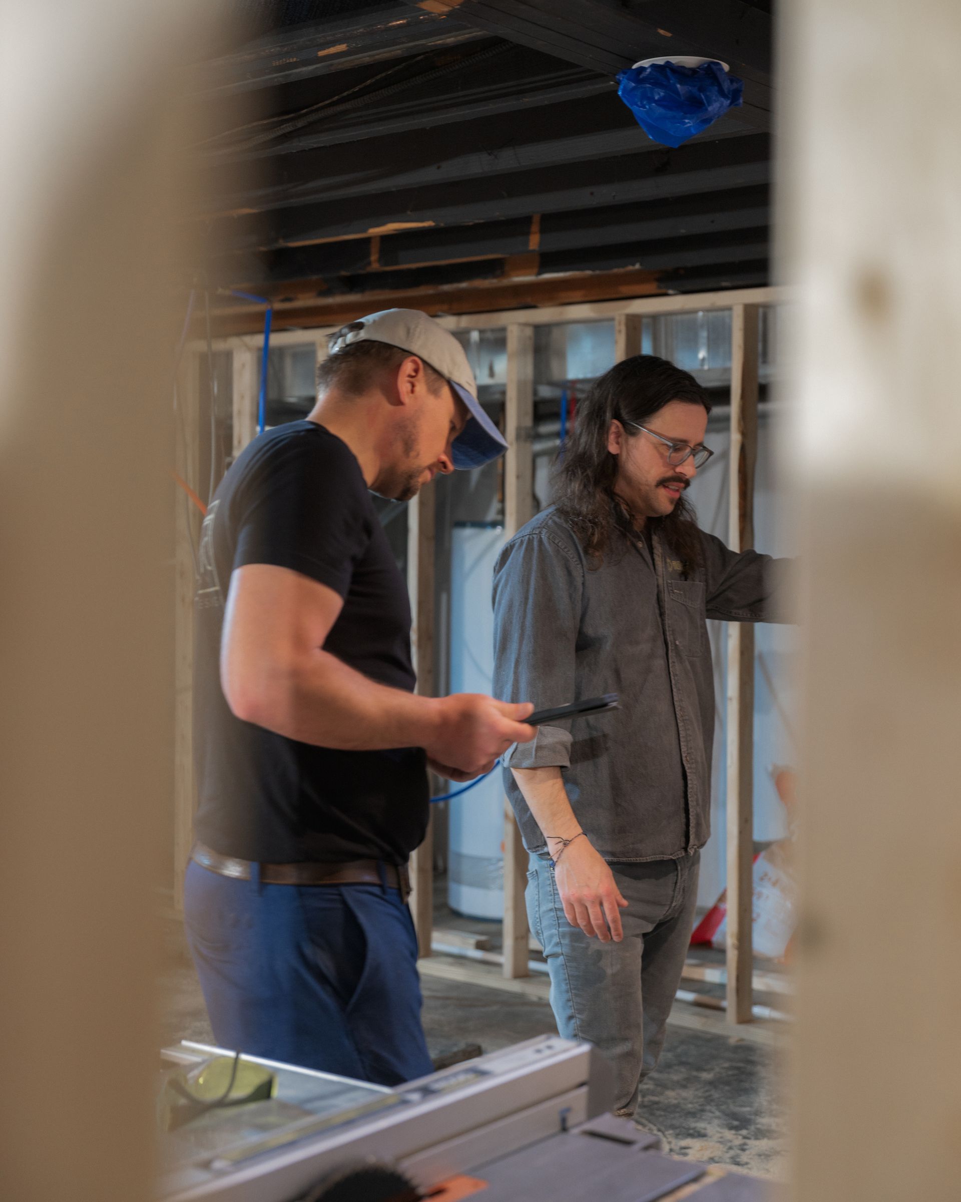 Two people in a partially built room inspect construction plans and framing.